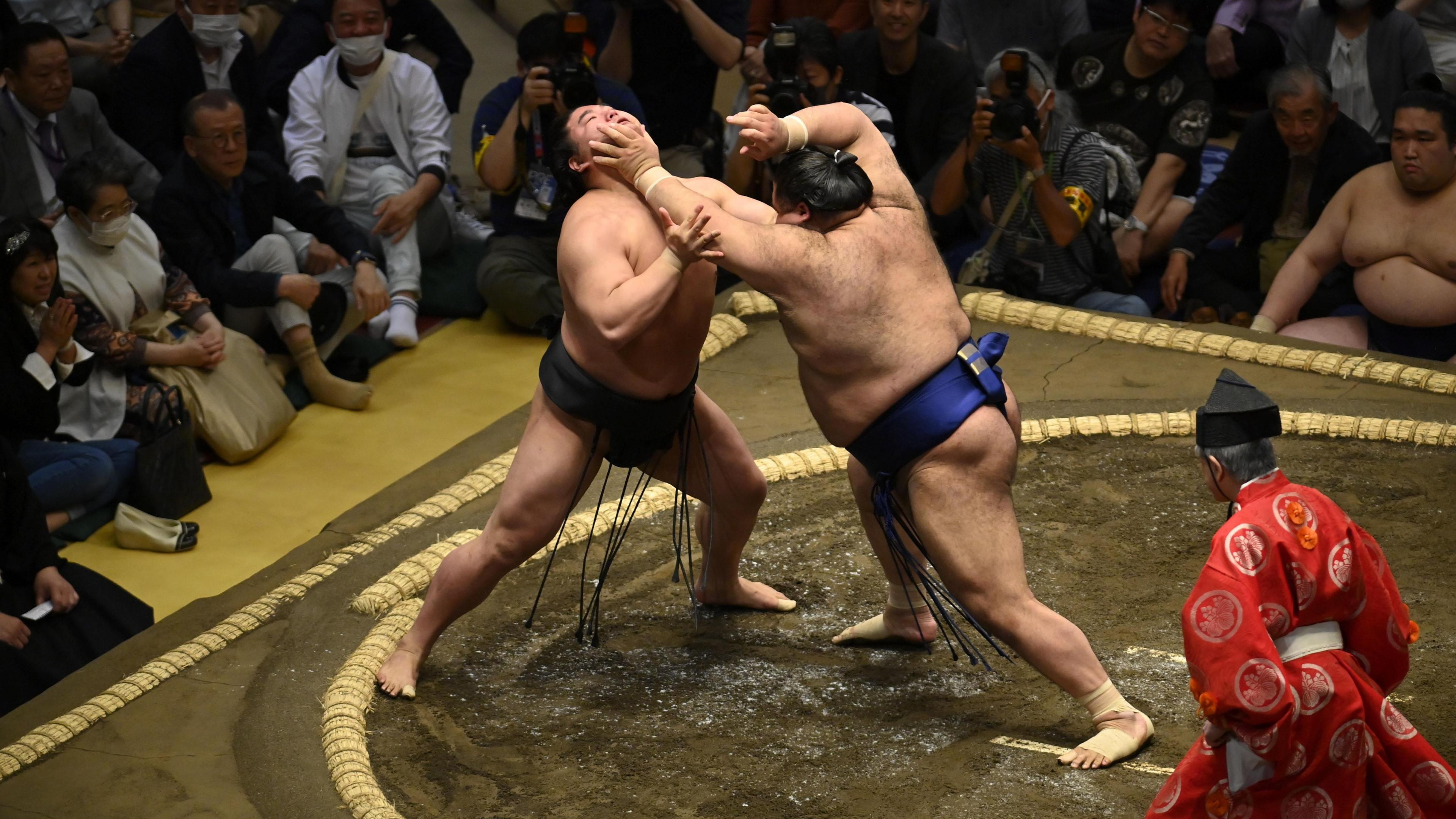 Two sumo wrestlers fight in the dohyo, while a red-dressed referee observes.
