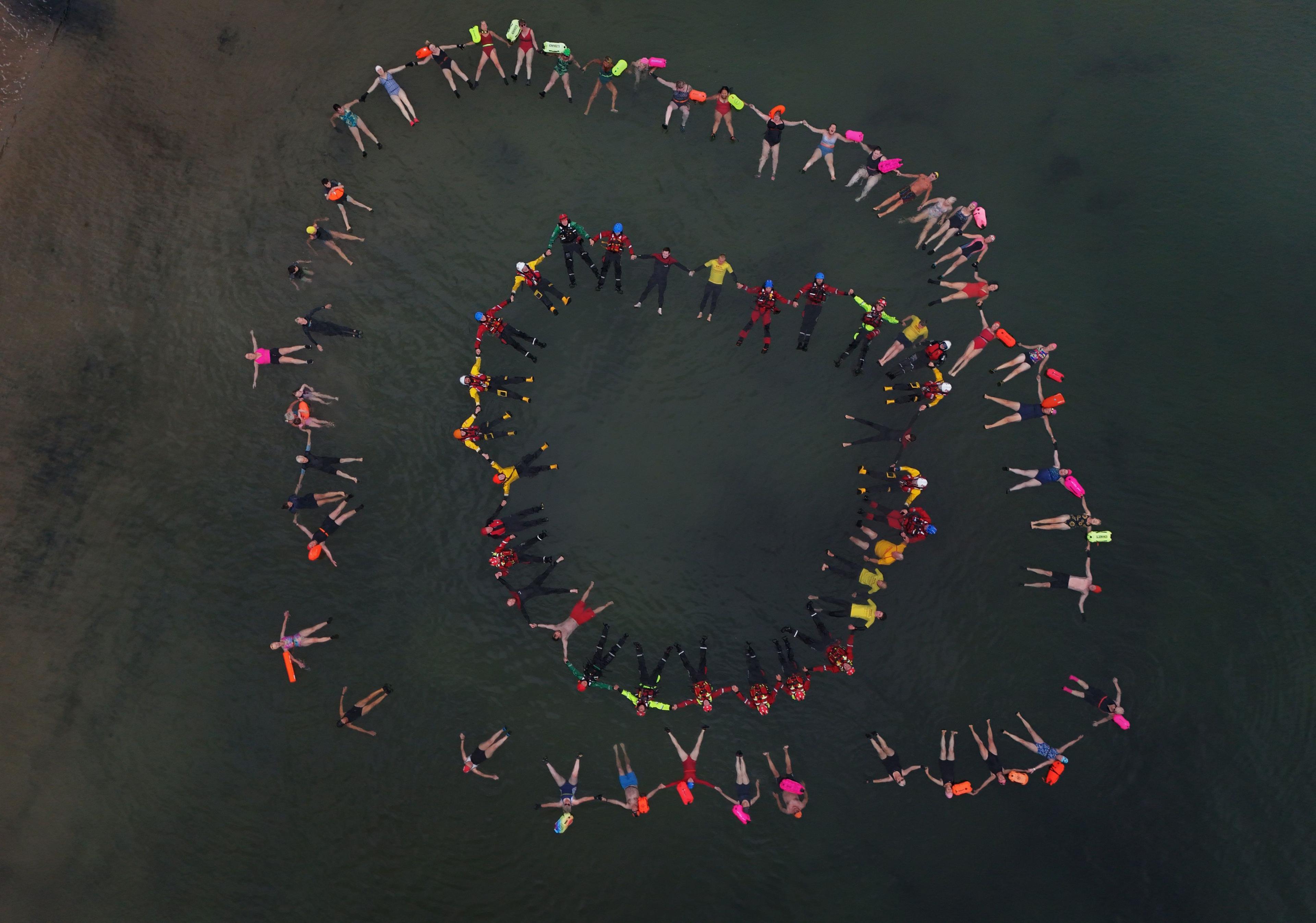 a group of swimmers floating together in a circle photographed from the air many are wearing brightly coloured clothes and holding hands