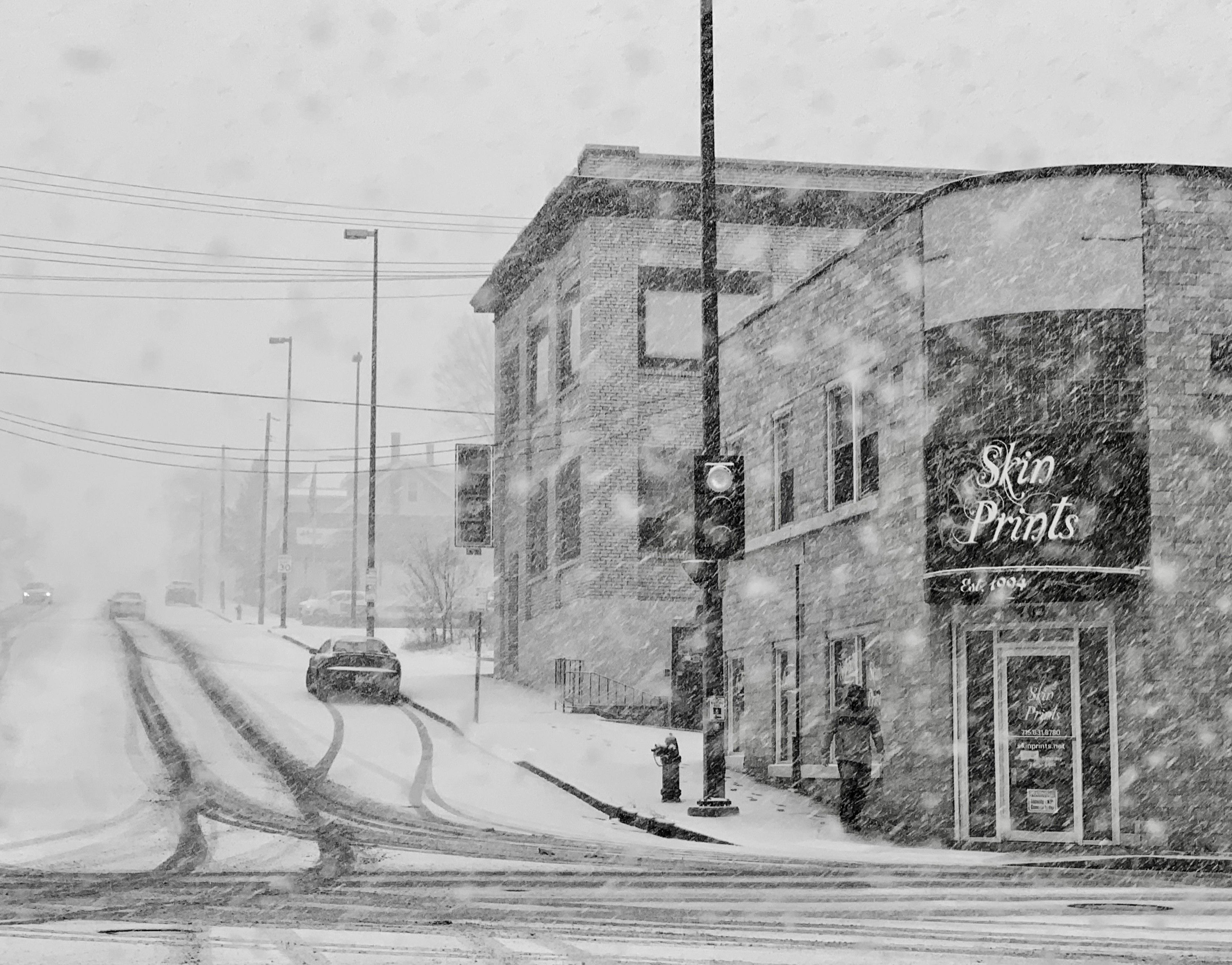 A black and white image of a road junction covered in snow. There is a shop on the right called 'Skin Prints' and cars driving up a snow-covered road on the left. A man is walking down the steep road, near the shop.