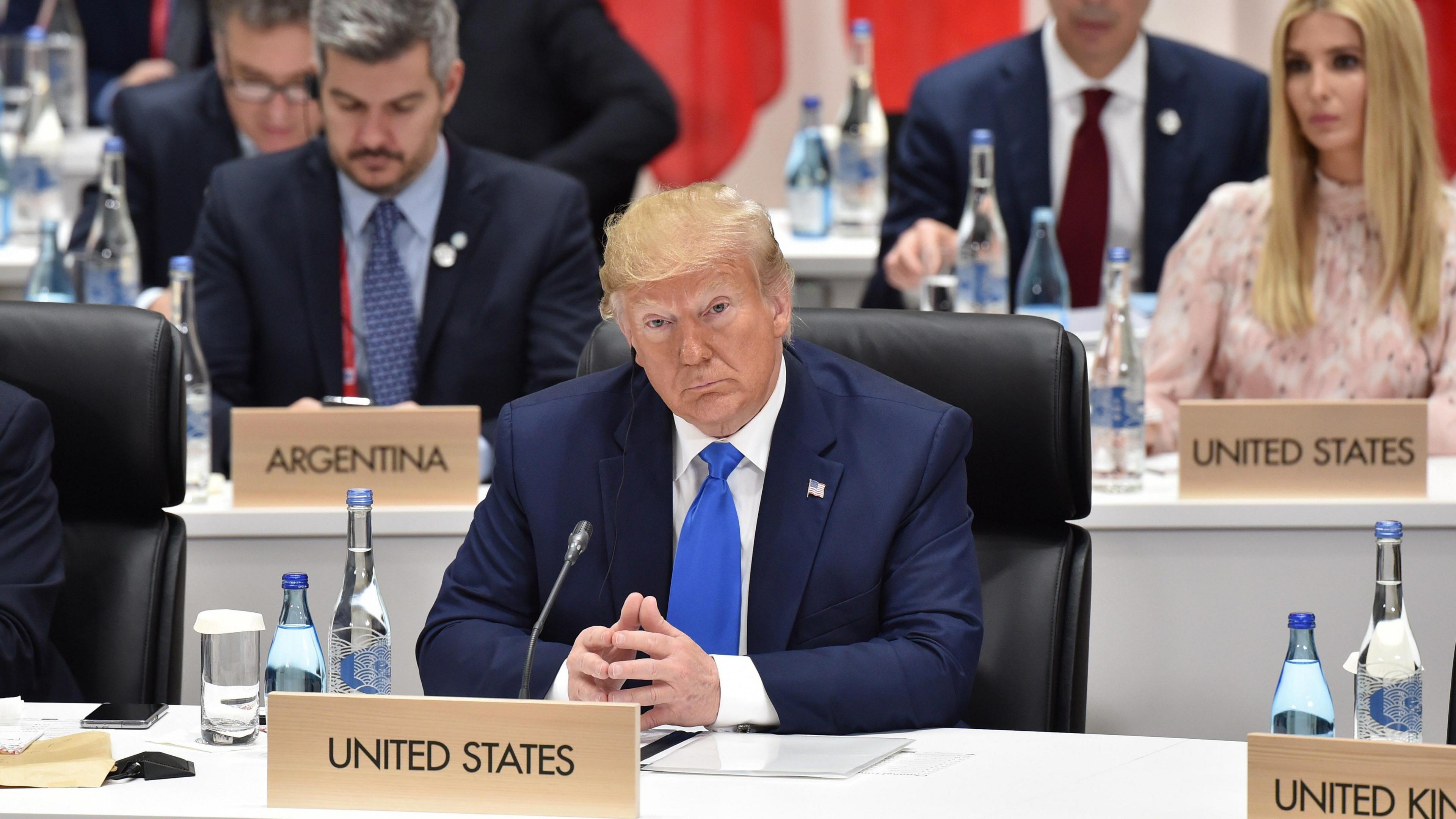 Donald Trump attends the G20 Summit in Osaka, Japan in 2019. He sits behind a United States sign, with his fingertips touching, and wears a blue suit with a blue tie. His daughter Ivanka Trump is visible at a desk behind his right shoulder. She has long blonde hair and wears a pale pink long-sleeved dress. 