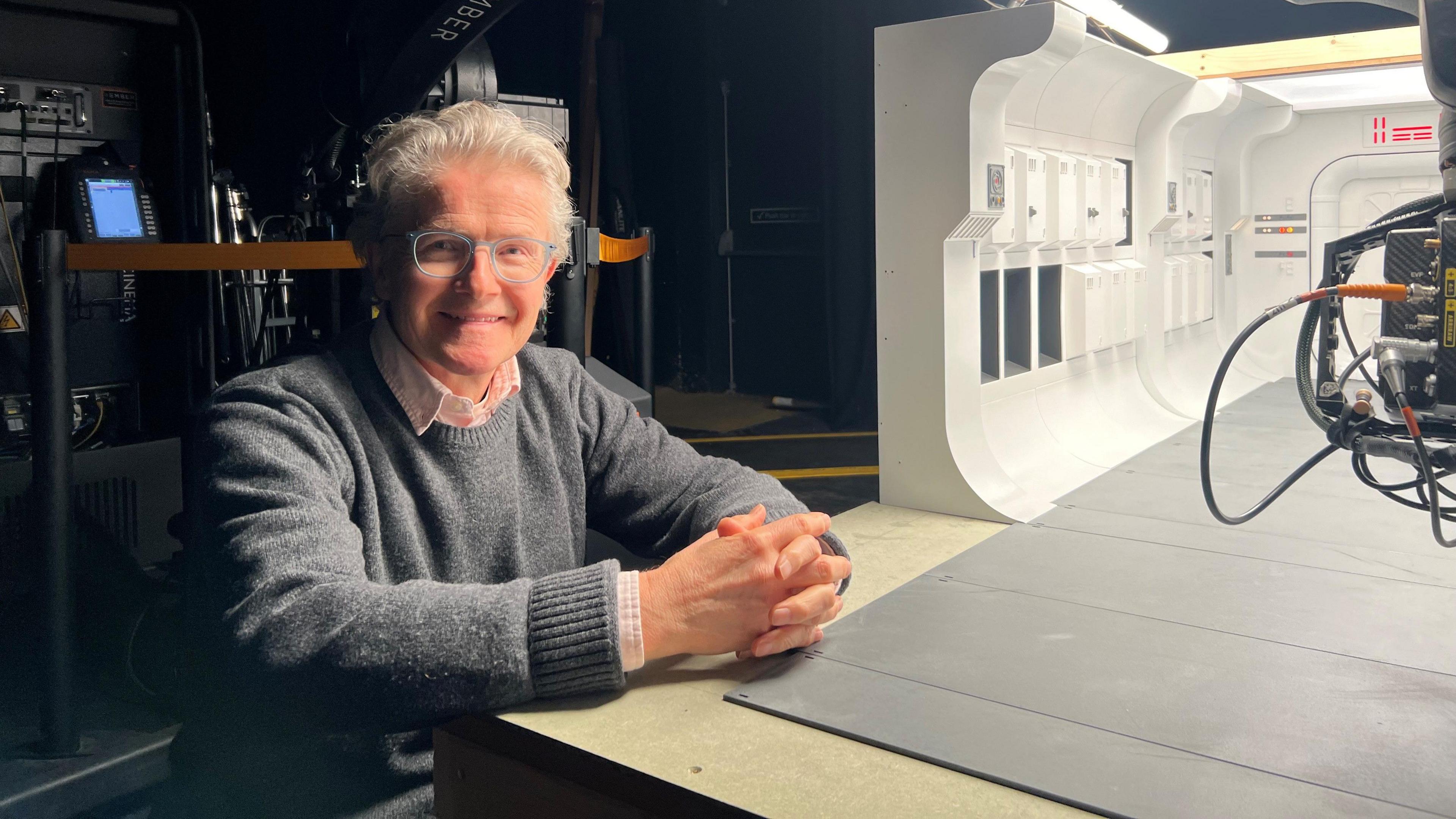John Lee is smiling, with silver hair and glasses, while stood near a model of a spaceship corridor set from Rogue One.