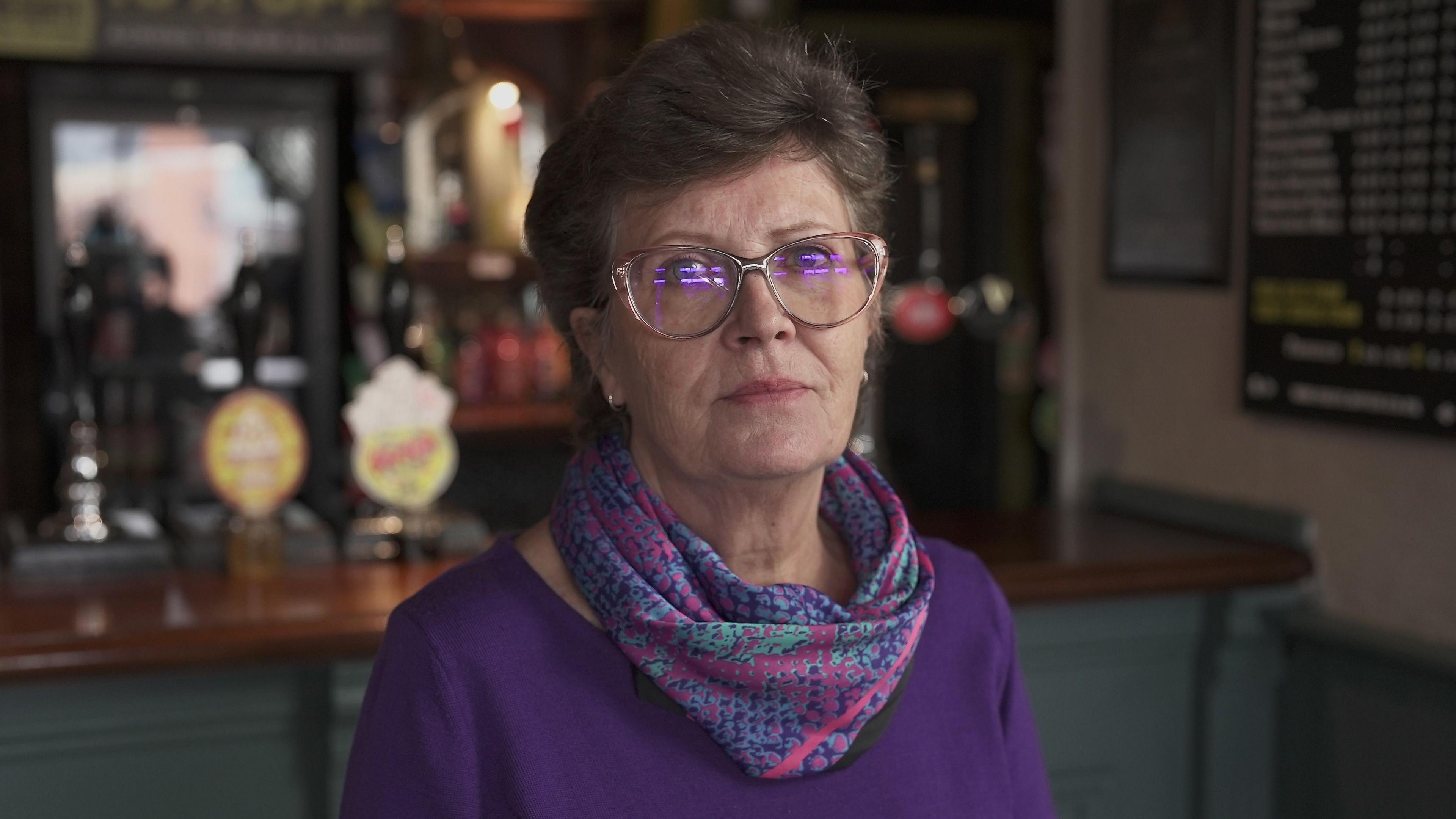 A woman in a purple top, with purple, pink and turquoise snake-skin print scarf. She has short hair and glasses and is looking at the camera with a pensive expression. Behind her a bar and beer taps can be seen.