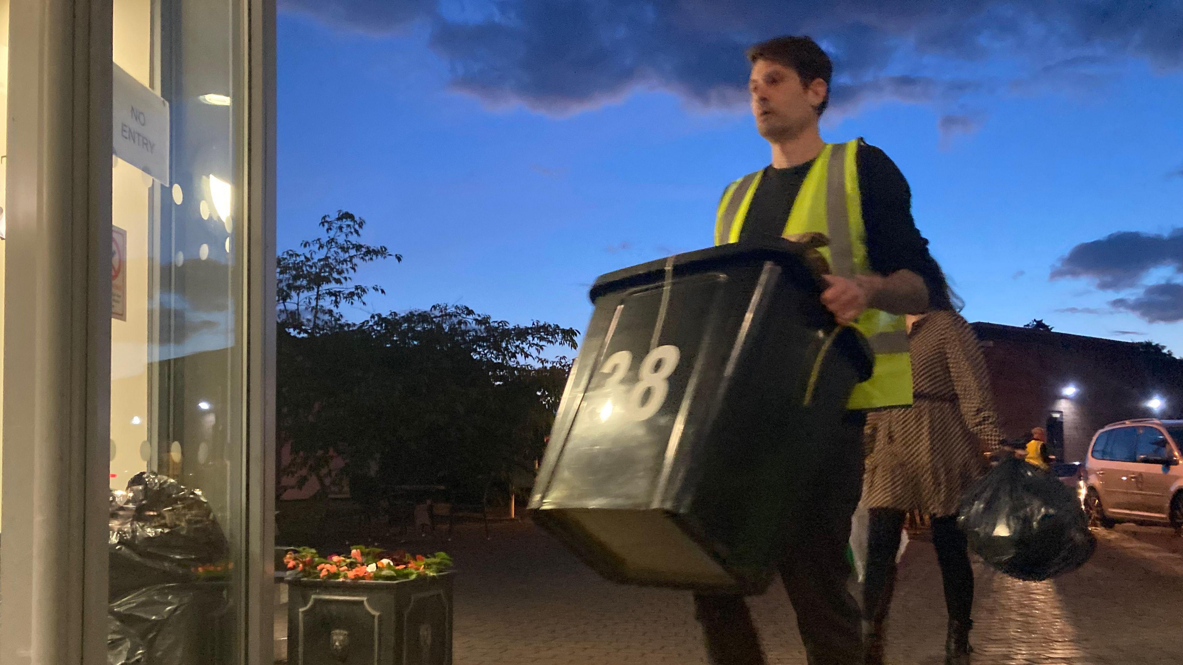 Aman in a hi vis vest carries a large plastic box towards a doorway a woman follows him with a black refuse sack