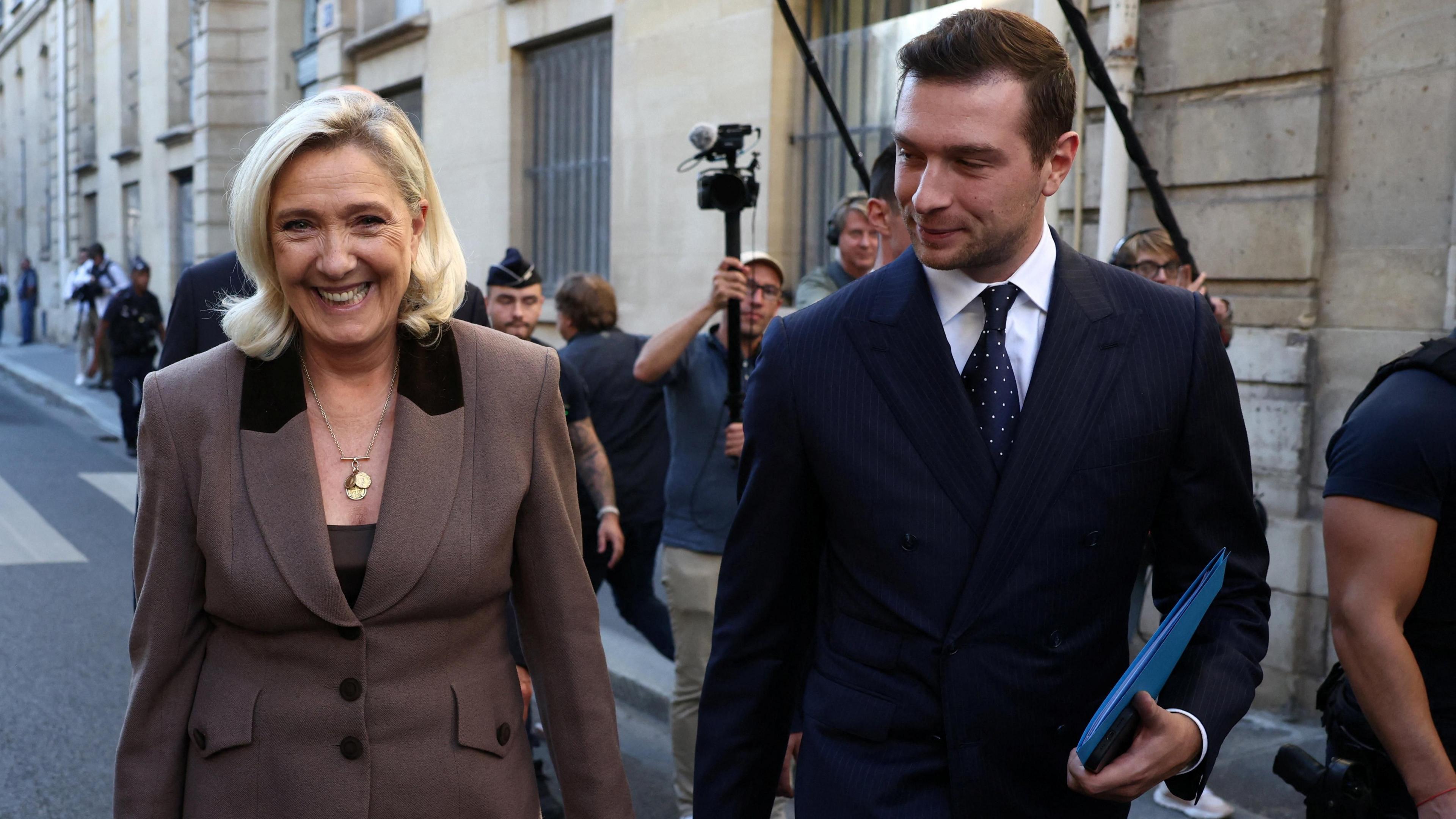 A smiling Marine Le Pen and Jordan Bardella walk down the street