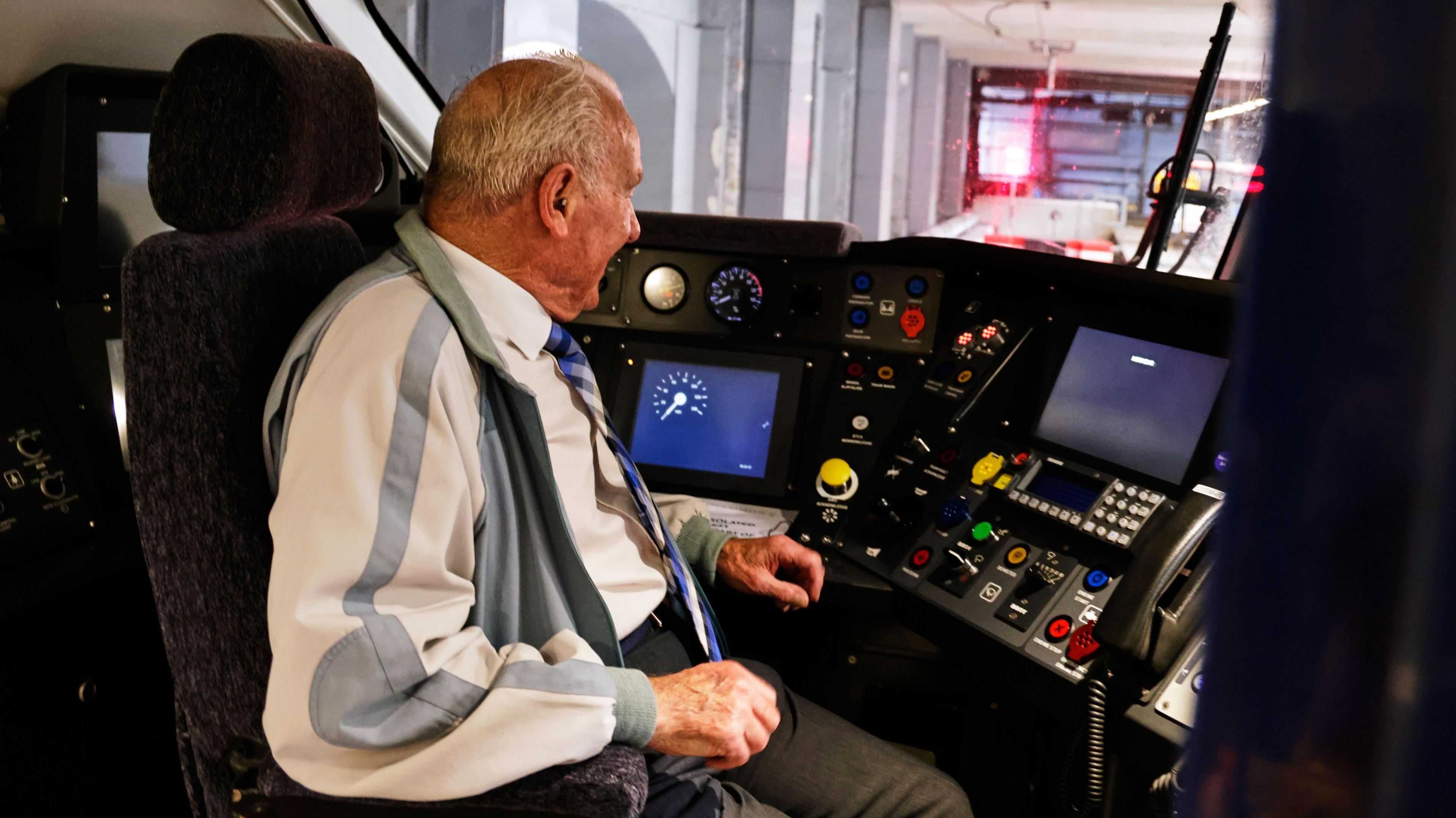 A man with short white hair, a white shirt, a blue patterned tie, dark trousers and a beige and grey jacket sitting in the cab of a train.