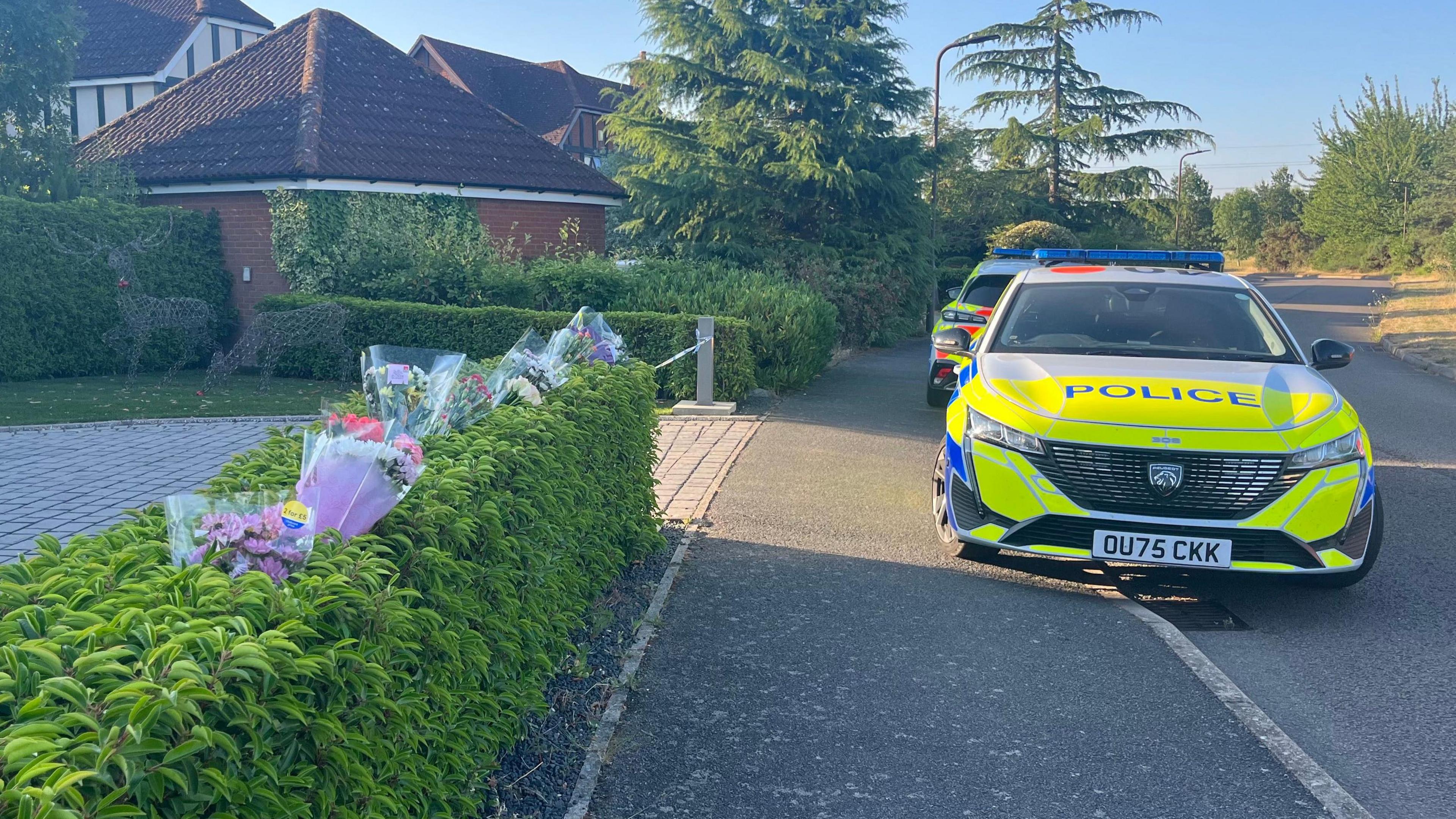 The scene at Carnoustie Drive in Great Denham. On the left is a hedge which has plastic-wrapped flowers sticking into it, and beyond is an entrance to a driveway with police tape across it. On the right are two police cars parked partly on the pavement. Beyond it is another detached house and trees.