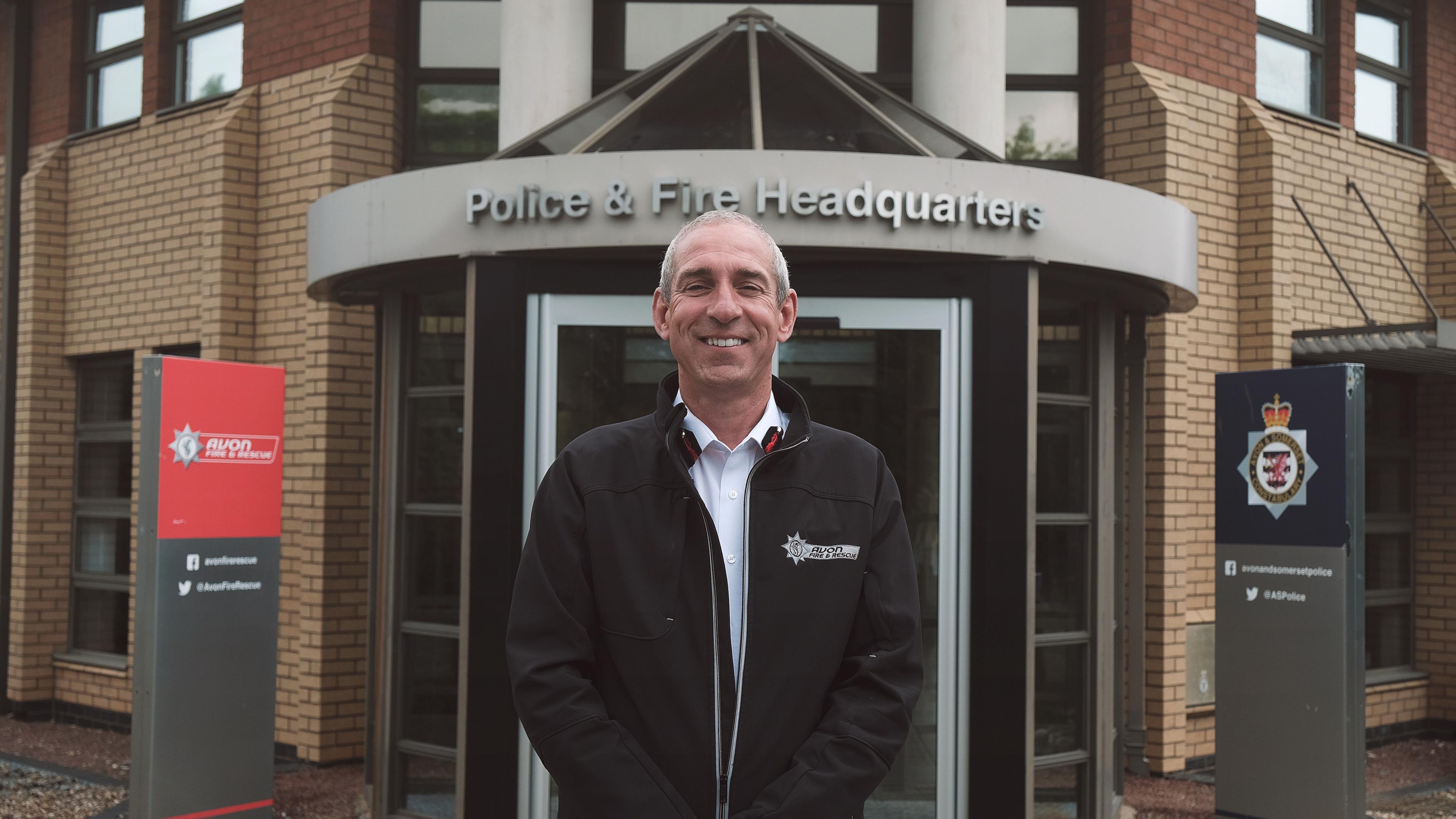 Simon Shilton pictured outside the fire and police HQ building. Simon has short grey hair and is wearing a black jacket with the Avon Fire and Rescue Service logo on the chest. He has a white shirt on underneath. He is standing with his hands in his pockets, looking directly at the camera and smiling. 