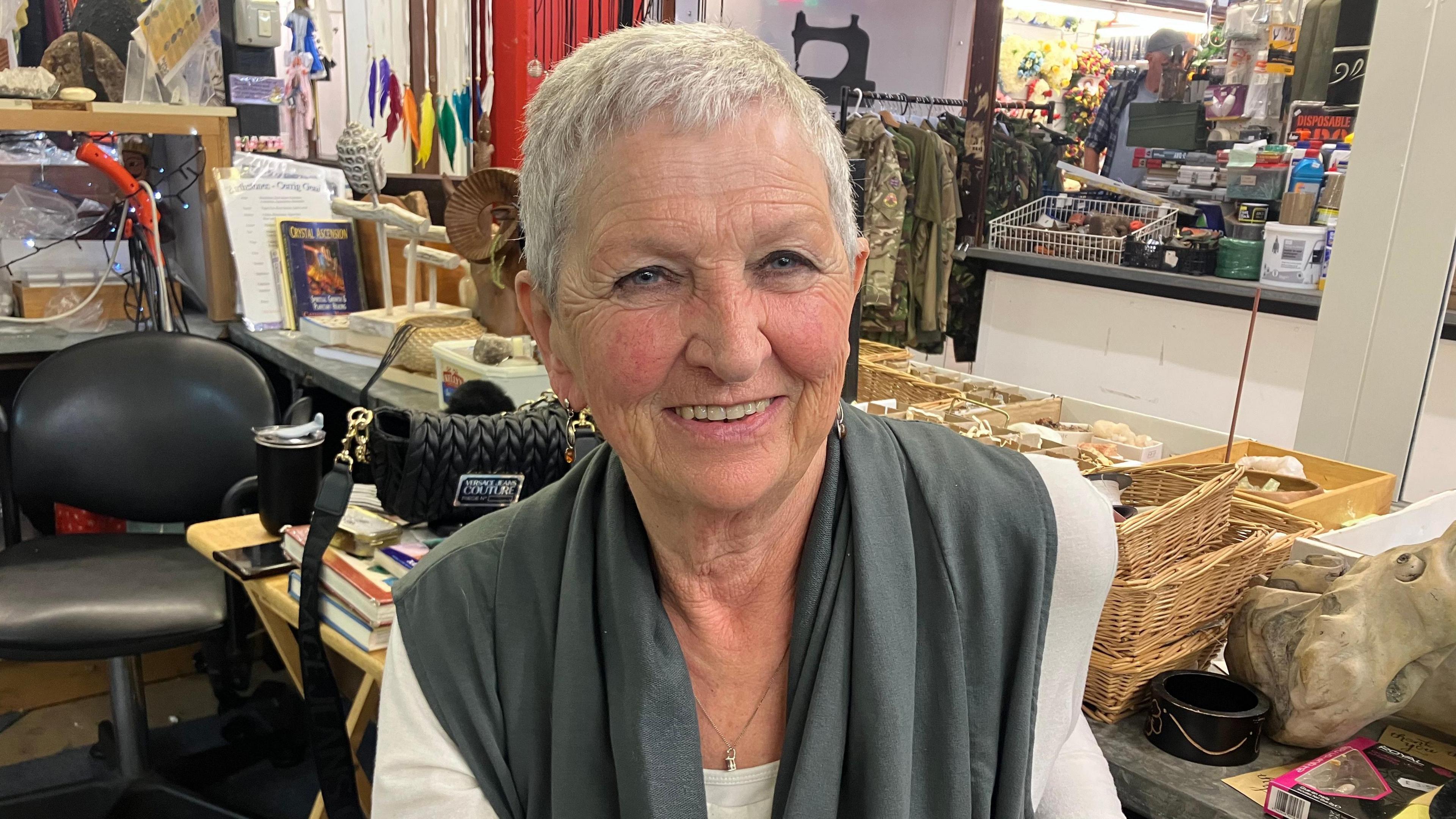 An older woman with short white hair, wear a long white sleeved top and a grey scarf