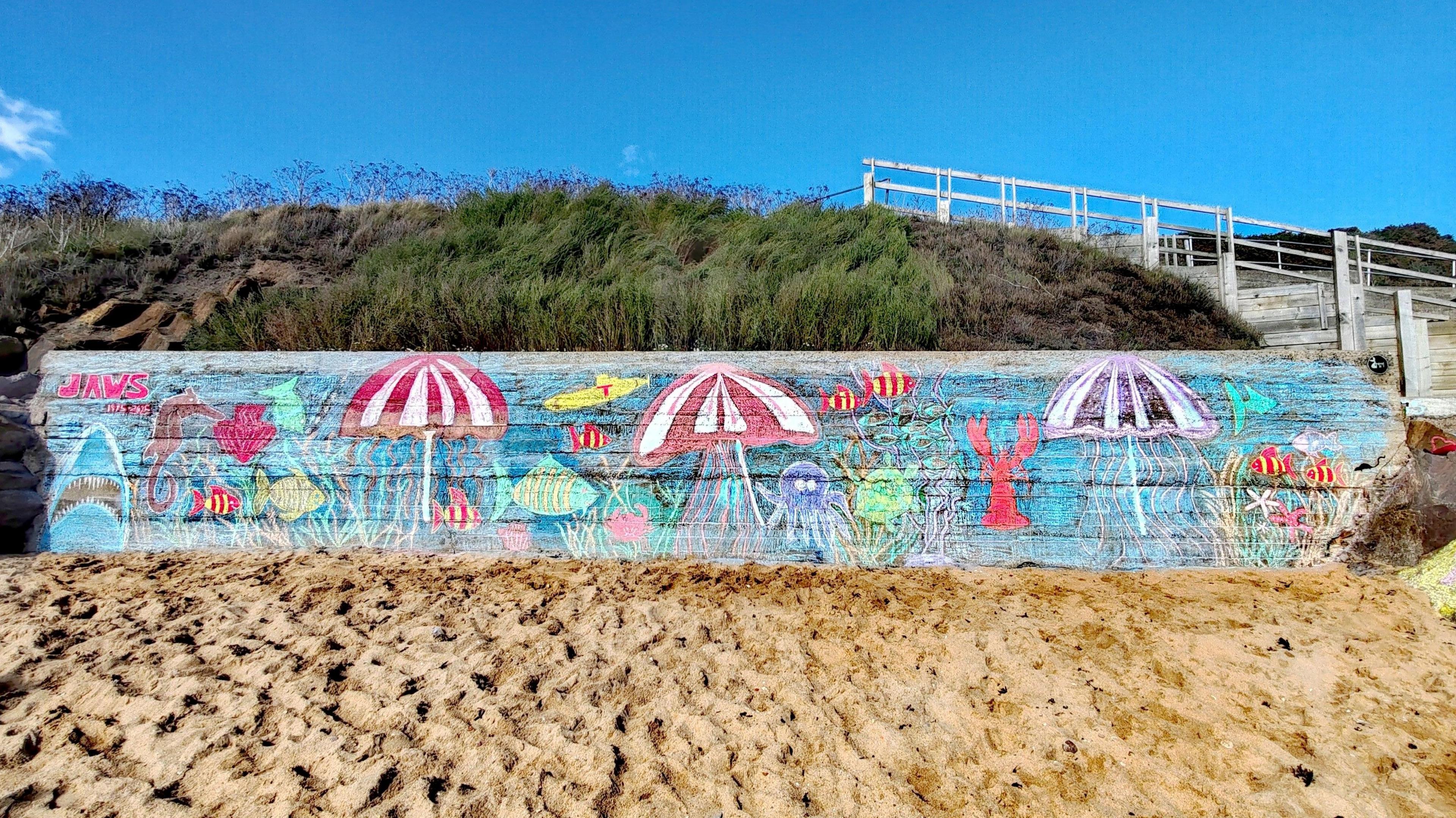 The chalk drawing on long piece of wood on a beach has a picture of Jaws the shark, an octopus, sun umbrellas, lobsters and fish set on a blue background.