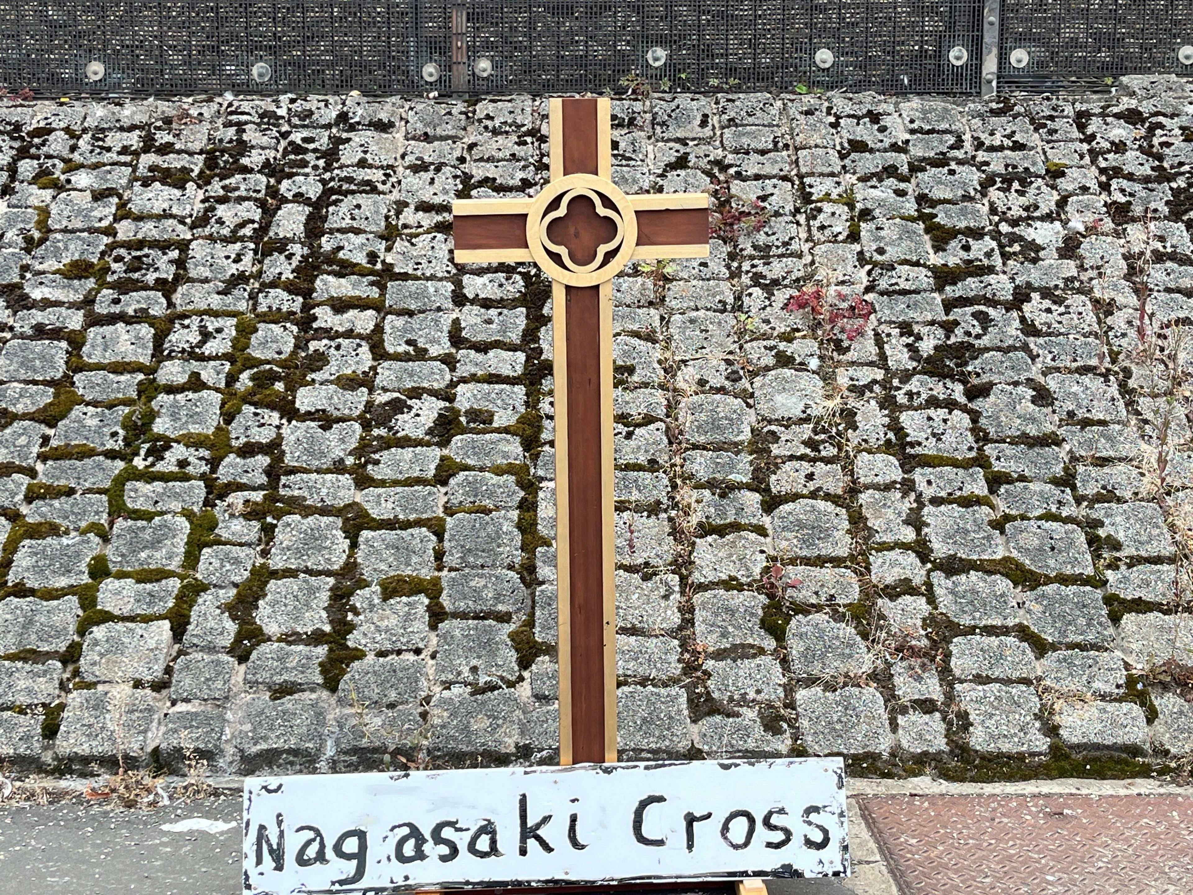 A wooden cross with a sign saying 'Nagasaki Cross' on a stone path, which is very mossy.