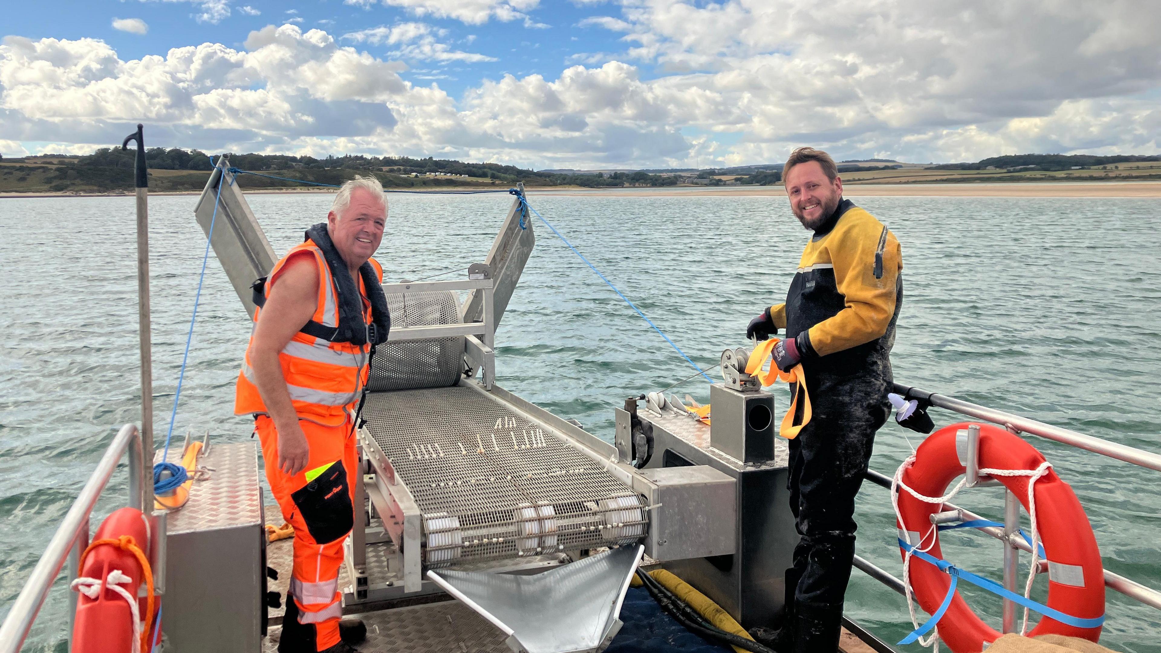 Two researchers from Life Wader standing on the deck of a boat. They are standing on either side of a conveyer bold at the rear of the vessel. They are both wearing wading gear. 