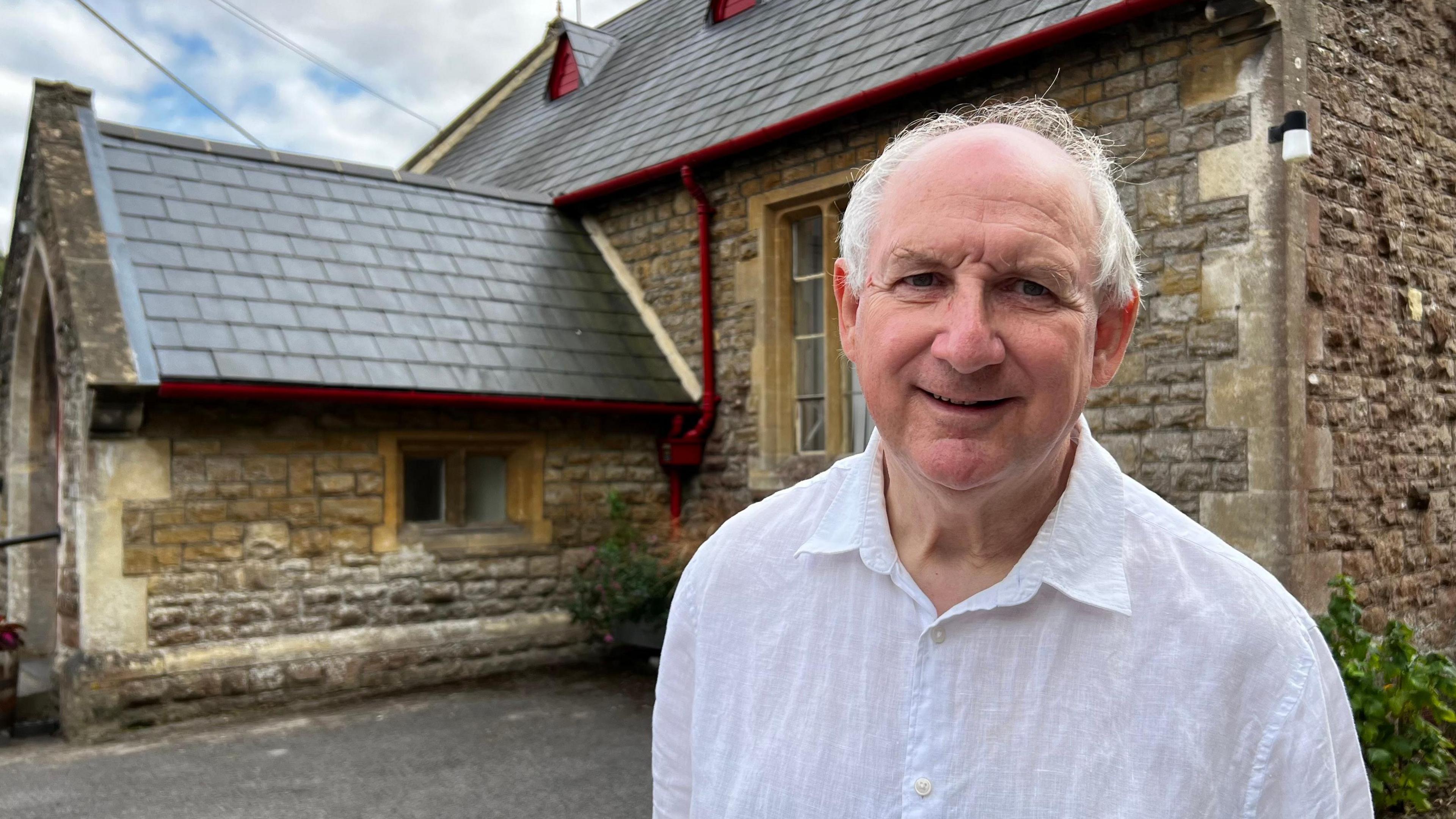 A man with white hair and wearing a white shirt stands outside a stone building with dark tiles on its roof. He is facing the camera and smiling