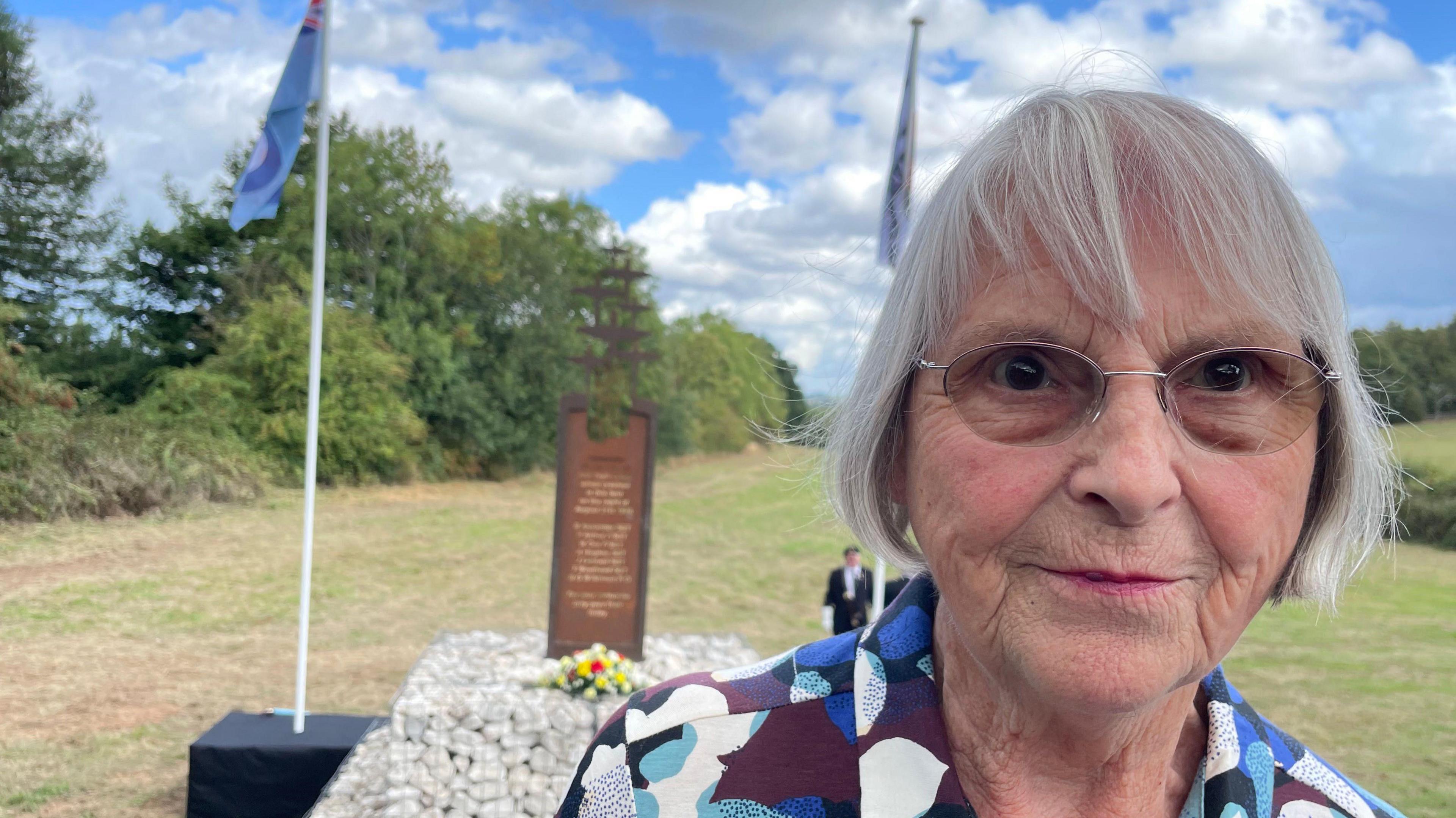 Mary Main, looking at the camera, with the memorial visible over her shoulder