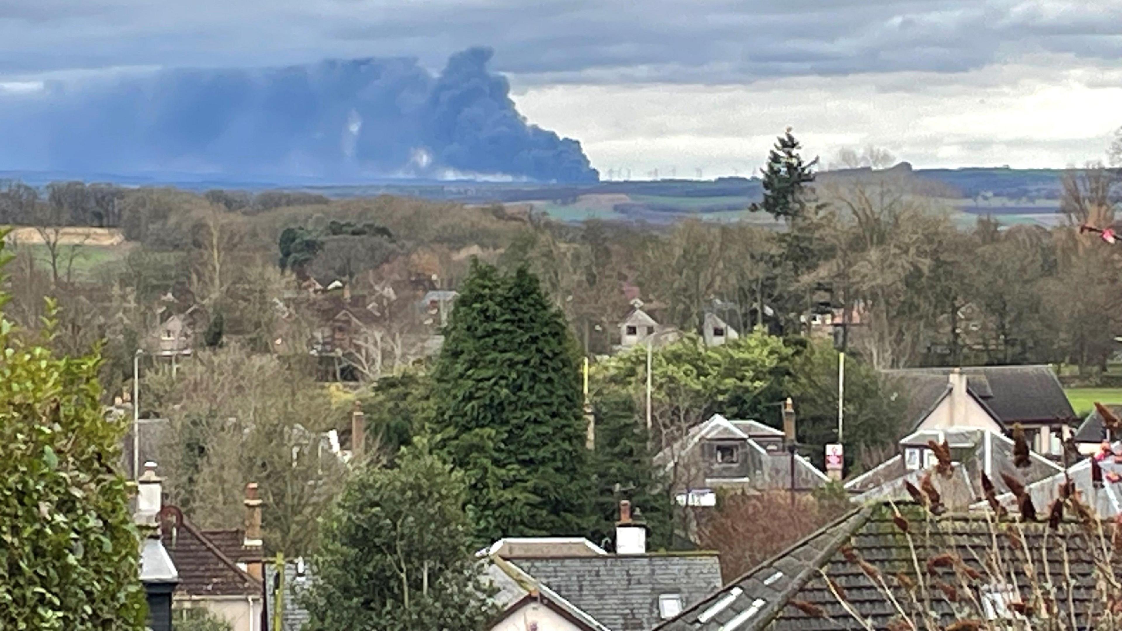 Smoke can be seen in the far distance from a high point 10 miles away in Fife. There are house roofs in the foreground and the smoke is rising in the distance near a wind farm on the horizon.