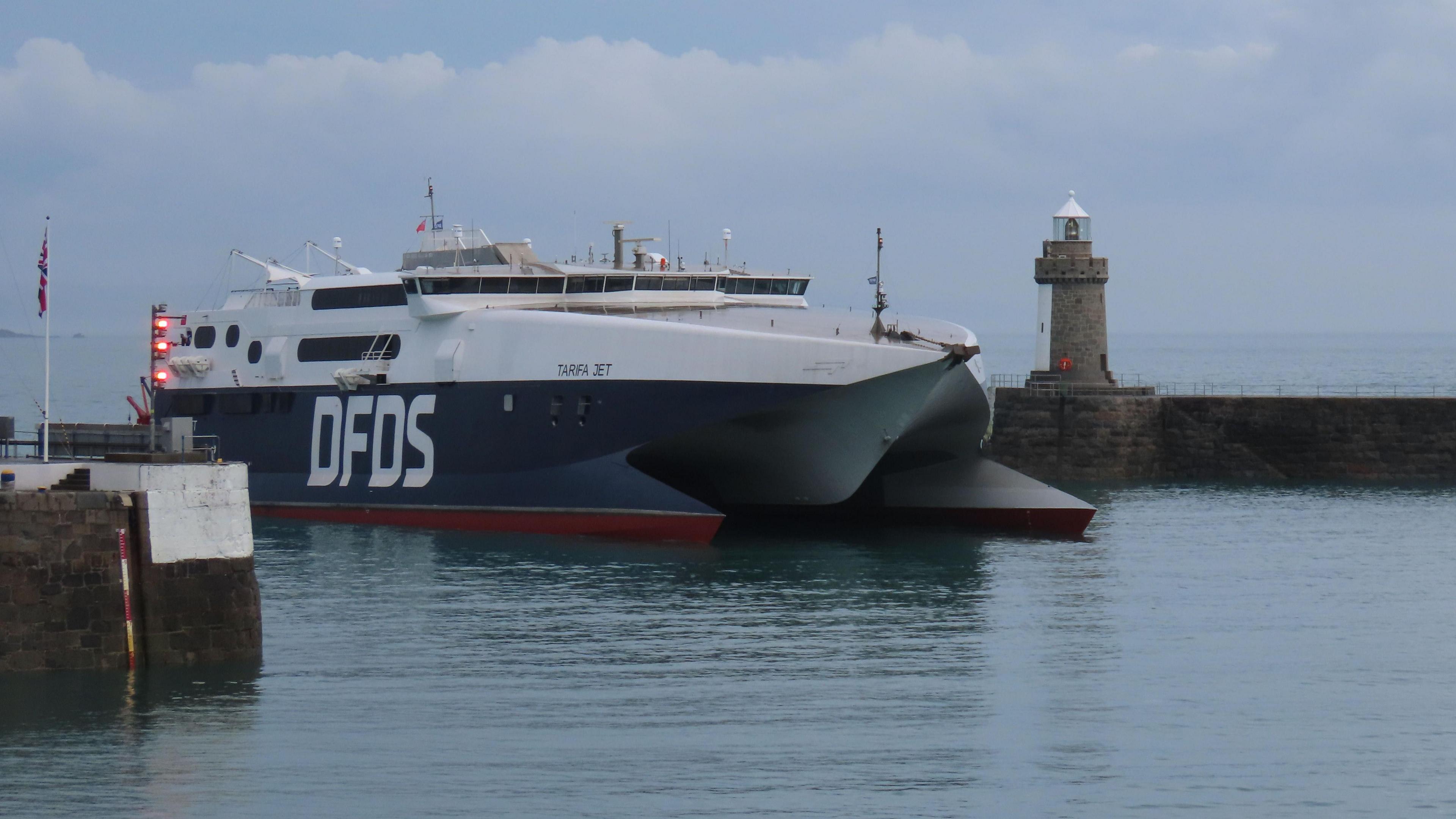 A wide shot of the DFDS coming into Guernsey Harbour. The sky is overcast. The boat is two tone, navy and white.