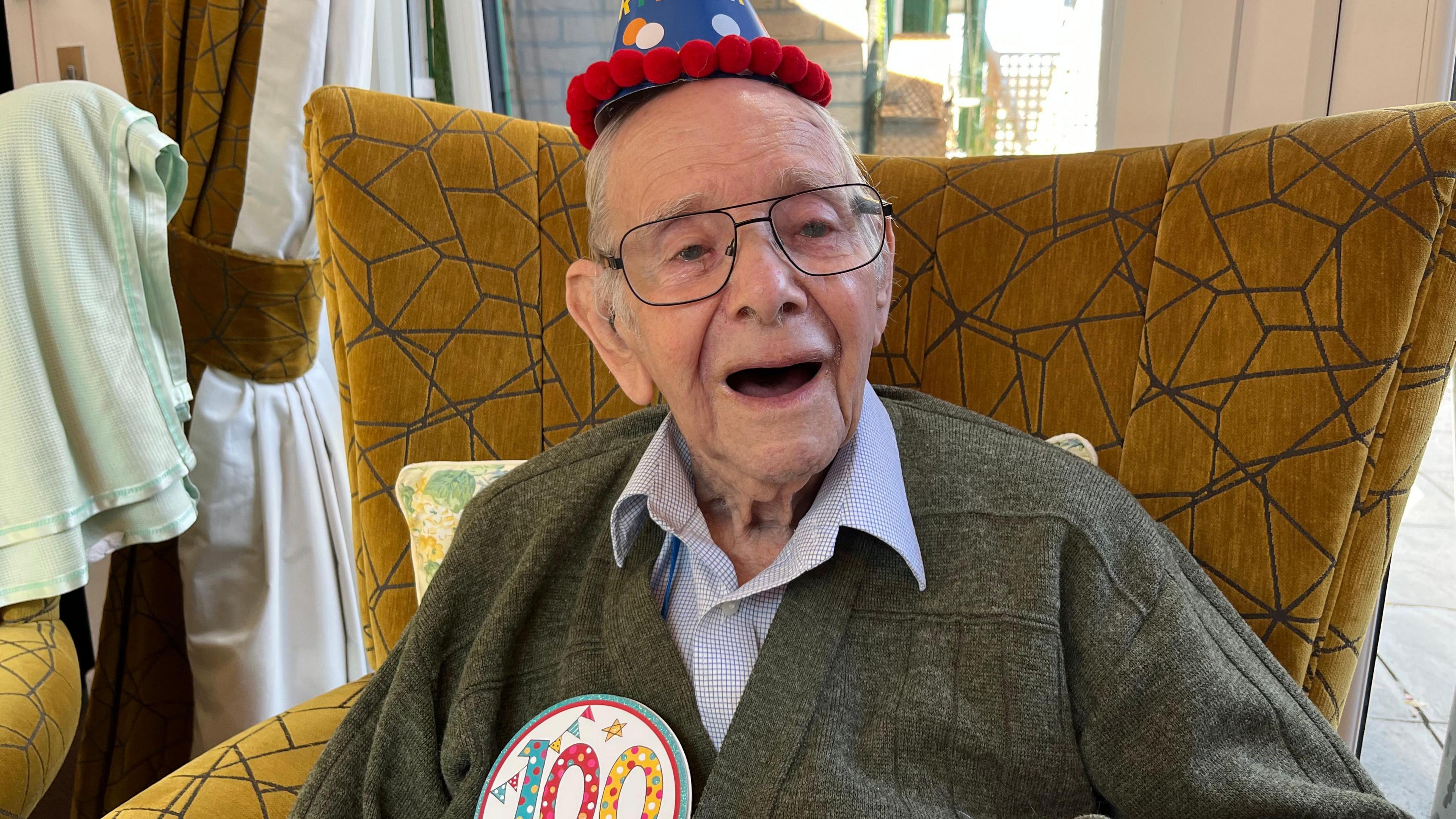Mr Cook is sat on a large brown chair while wearing a 100 badge and a blue and red party hat. He is smiling and also wears a pair or large framed glasses. He has on a blue shirt and grey cardigan.