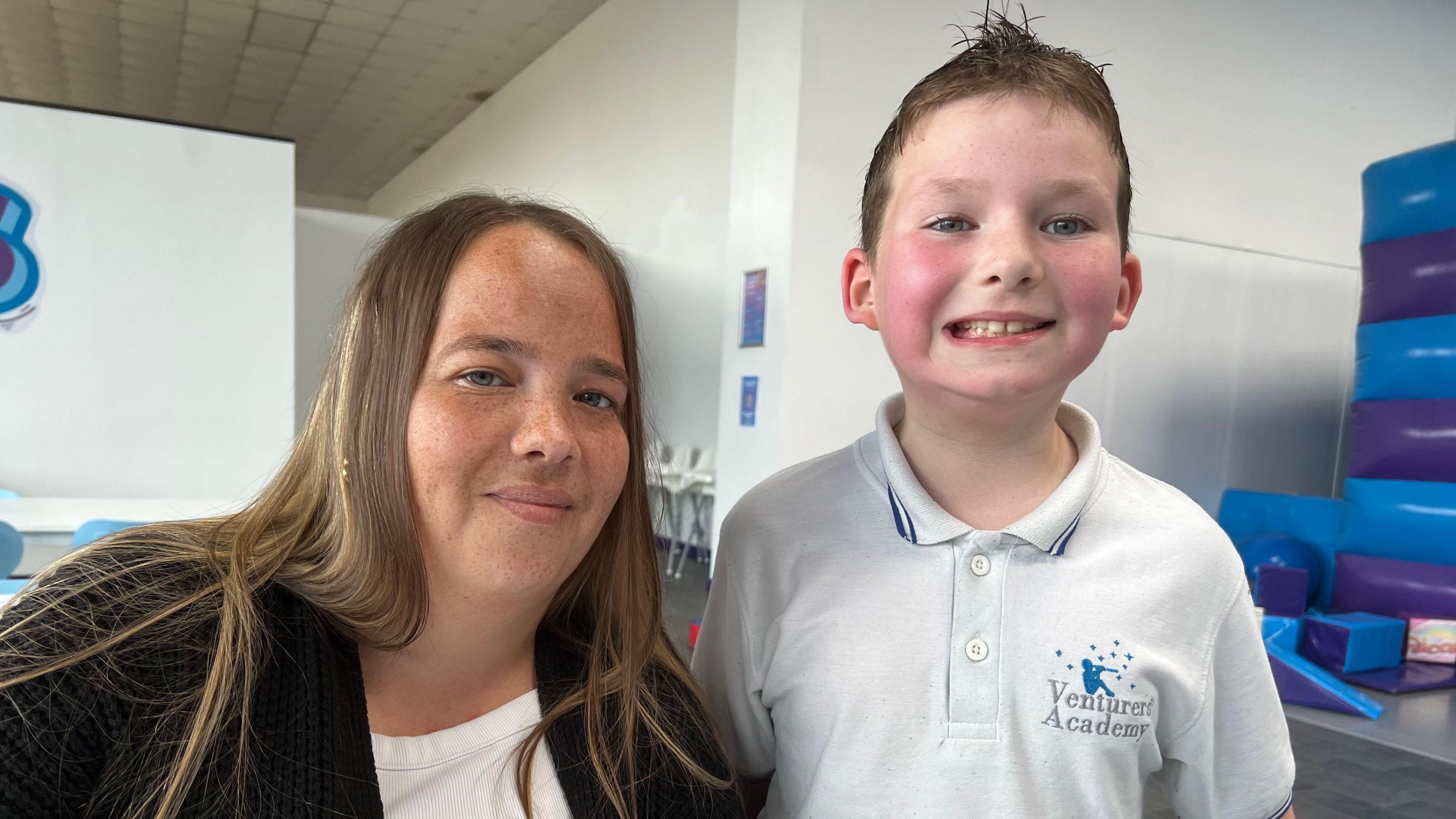 Trojeana wearing a white top and black cardigan. She has long and straight brown hair and is smiling at the camera. Beside her is Ted, grinning with pink cheeks and wearing a white polo shirt with buttons.