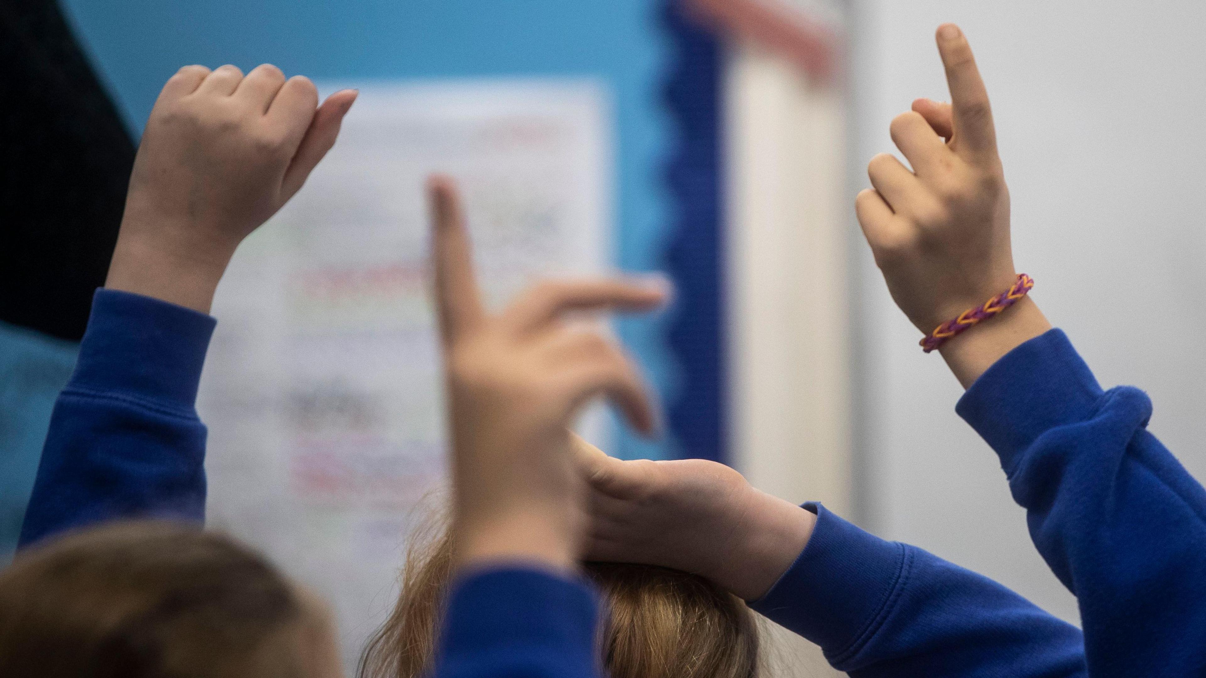 Four primary school children's hands held up in a classroom. All are wearing blue school sweatshirts. One is wearing a orange and purple bracelet.