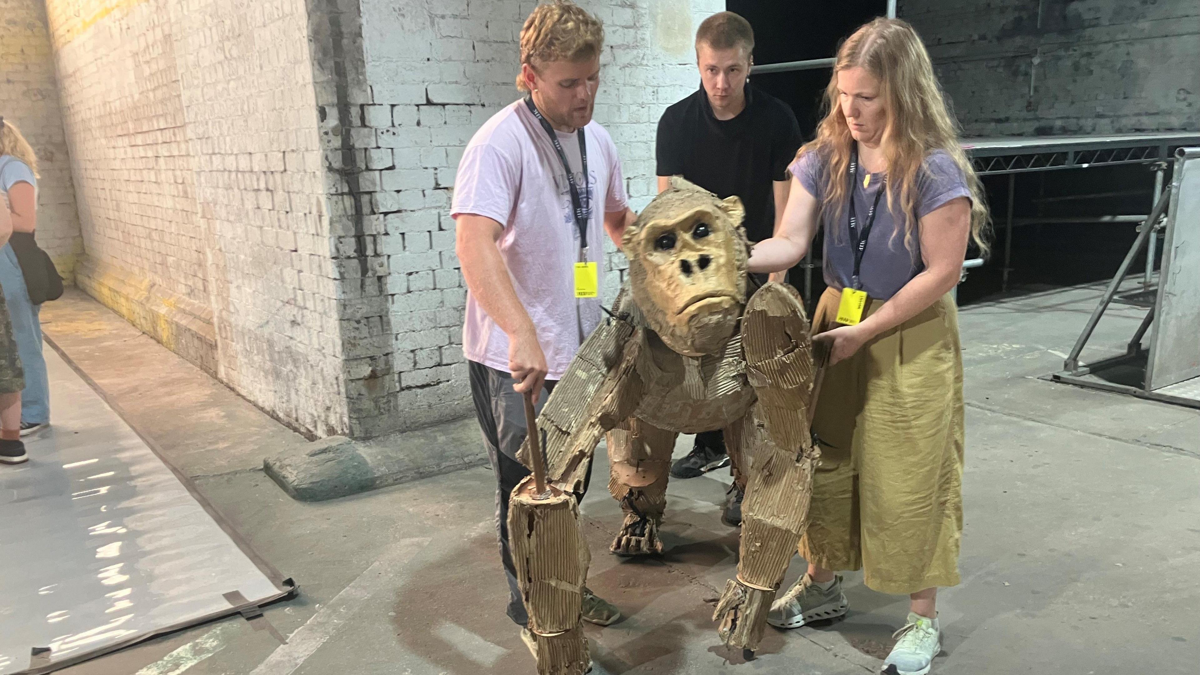 Photograph of three people controlling a puppet gorilla. The picture was taken inside a rehearsal space under railway arches in Manchester city centre.