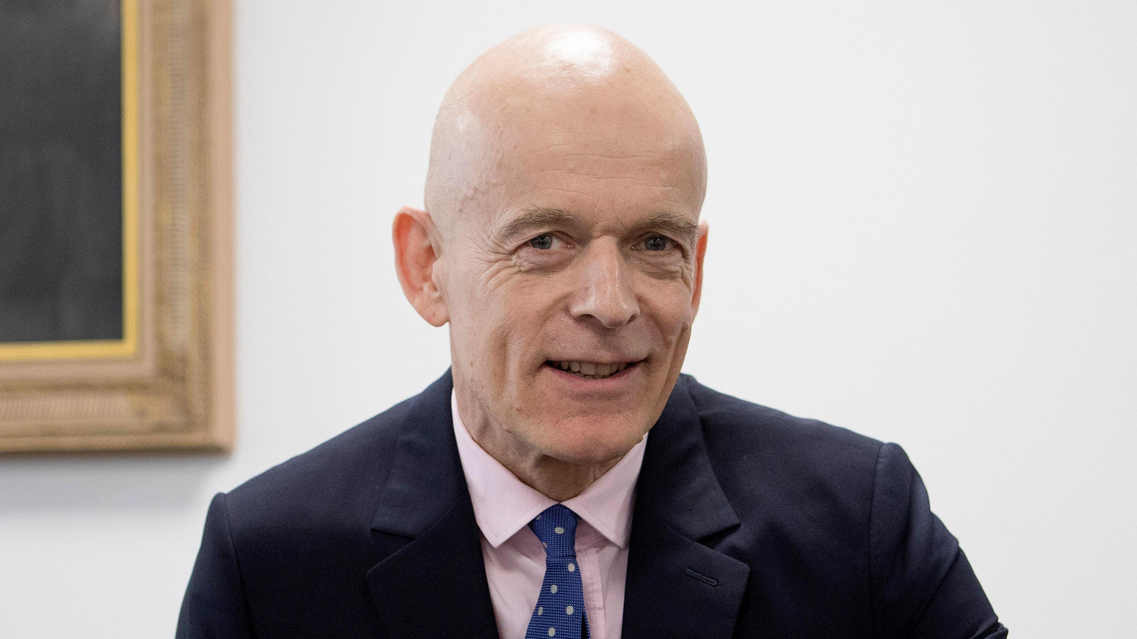 Sir Adrian Fulford, who has a bald head and is wearing a navy blue suit over a pink collared shirt, smiles at the camera