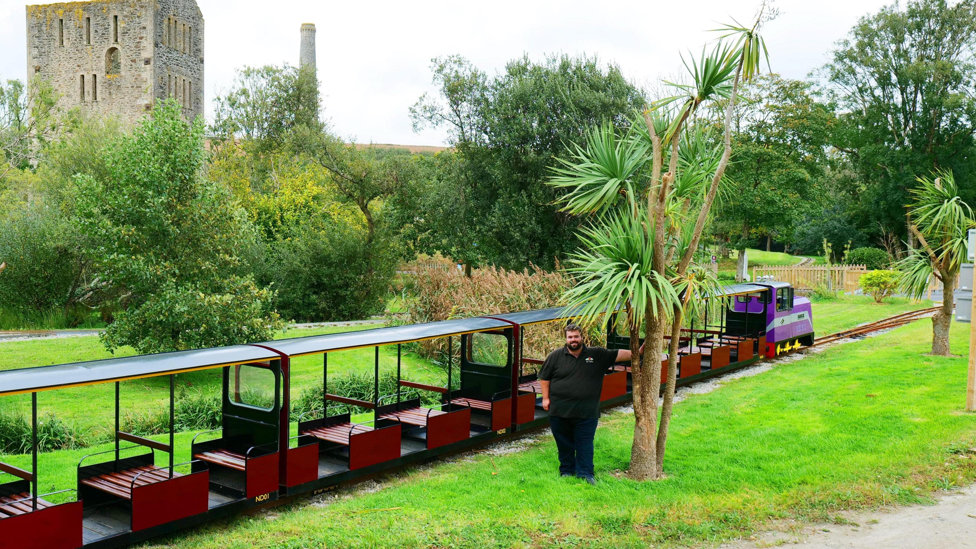 Ben Harding stood in by one of the newly planted palm trees. The trees are about three times his height. He is stood in the grounds of the tourist attraction, wearing black trousers and a black shirt. He has dark hair and a beard. Directly behind him is one of the trains and it's carriages. Each carriage has red painted wooden seats. The train is a bright purple colour. Behind the train there is grass, bushes a path and an old building which looks similar to a castle. 