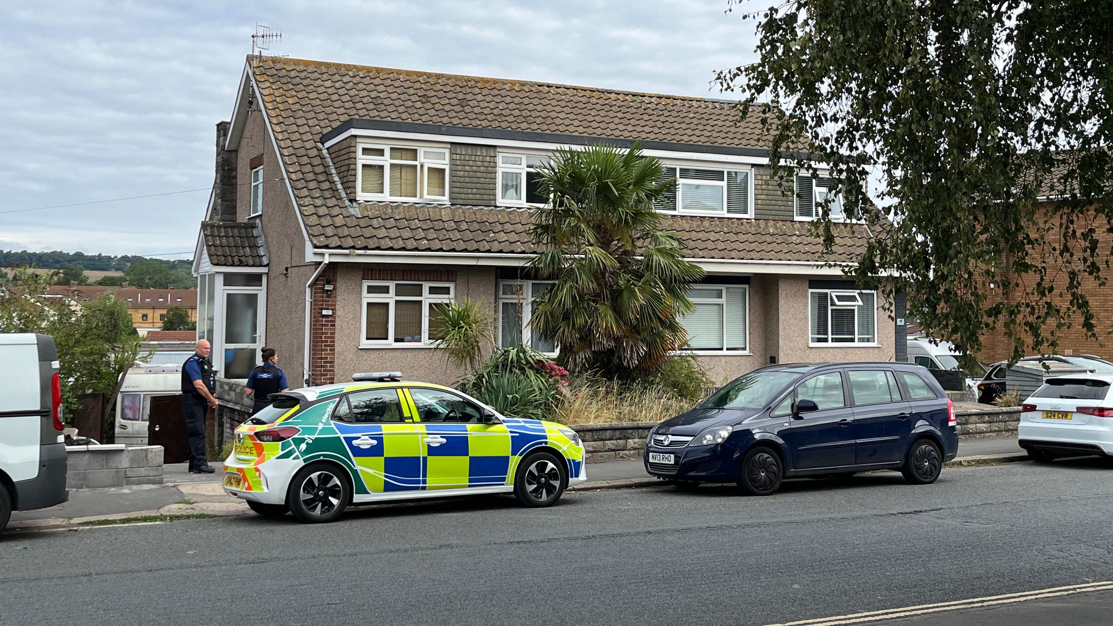 Two police officers in uniform standing outside a home in South Liberty Lane during the daytime, following the incident. It is a pebbledash building with slate roofs and a large palm tree outside. There are cars parked beside the pavement, including a police car with hi-vis blue and yellow decals.
