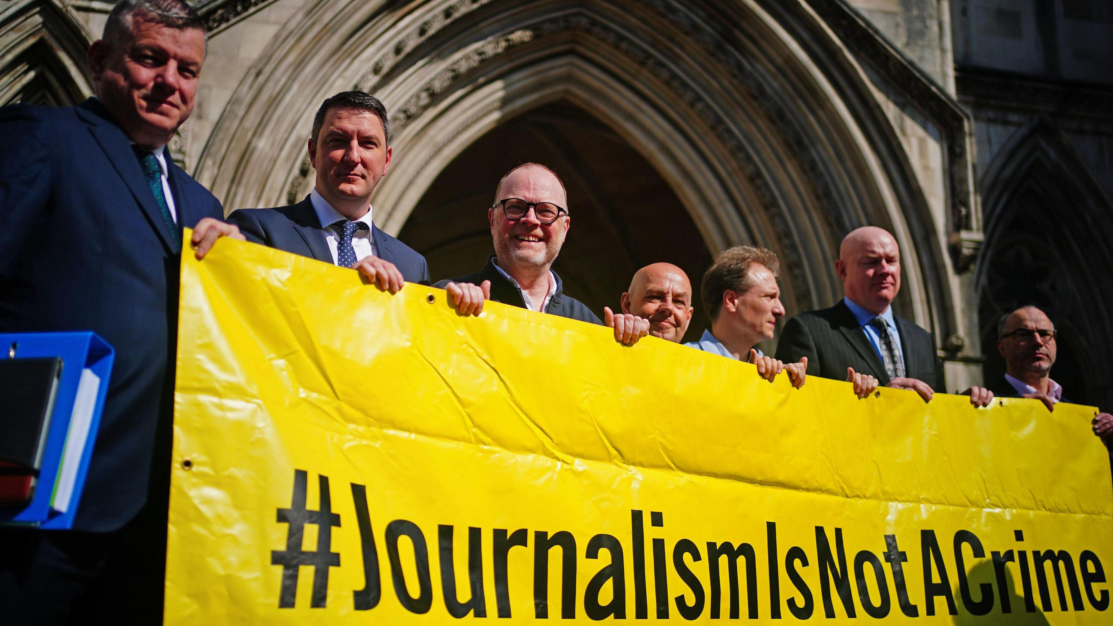 Supporters of Barry McCaffrey and Trevor Birney outside the Royal Courts of Justice in London. They are holding a yellow banner with black text that says "#JournalismIsNotACrime"