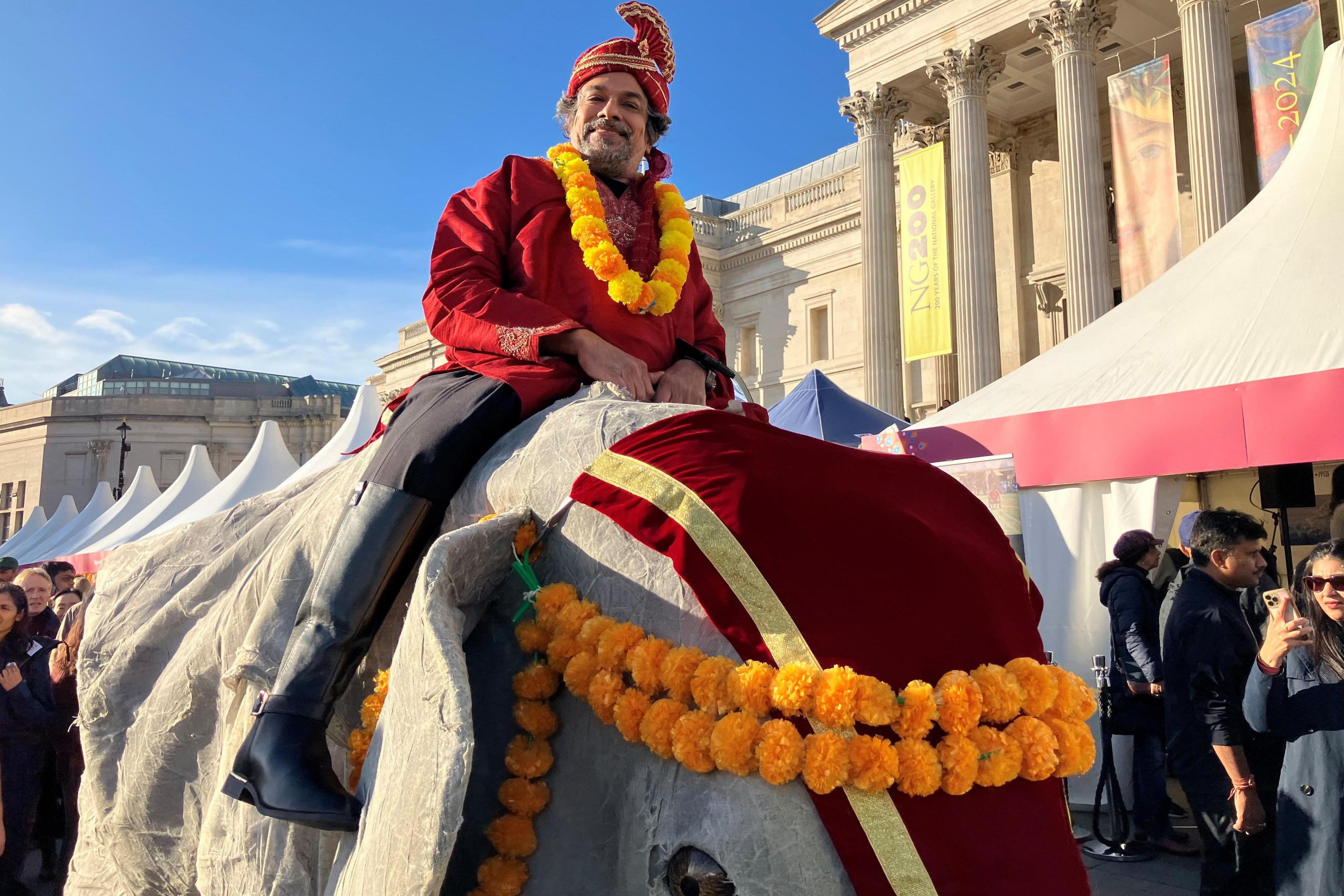 A man in a red turban and yellow garland wearing a comedy costume of an elephant, which makes him look like he is sitting on it