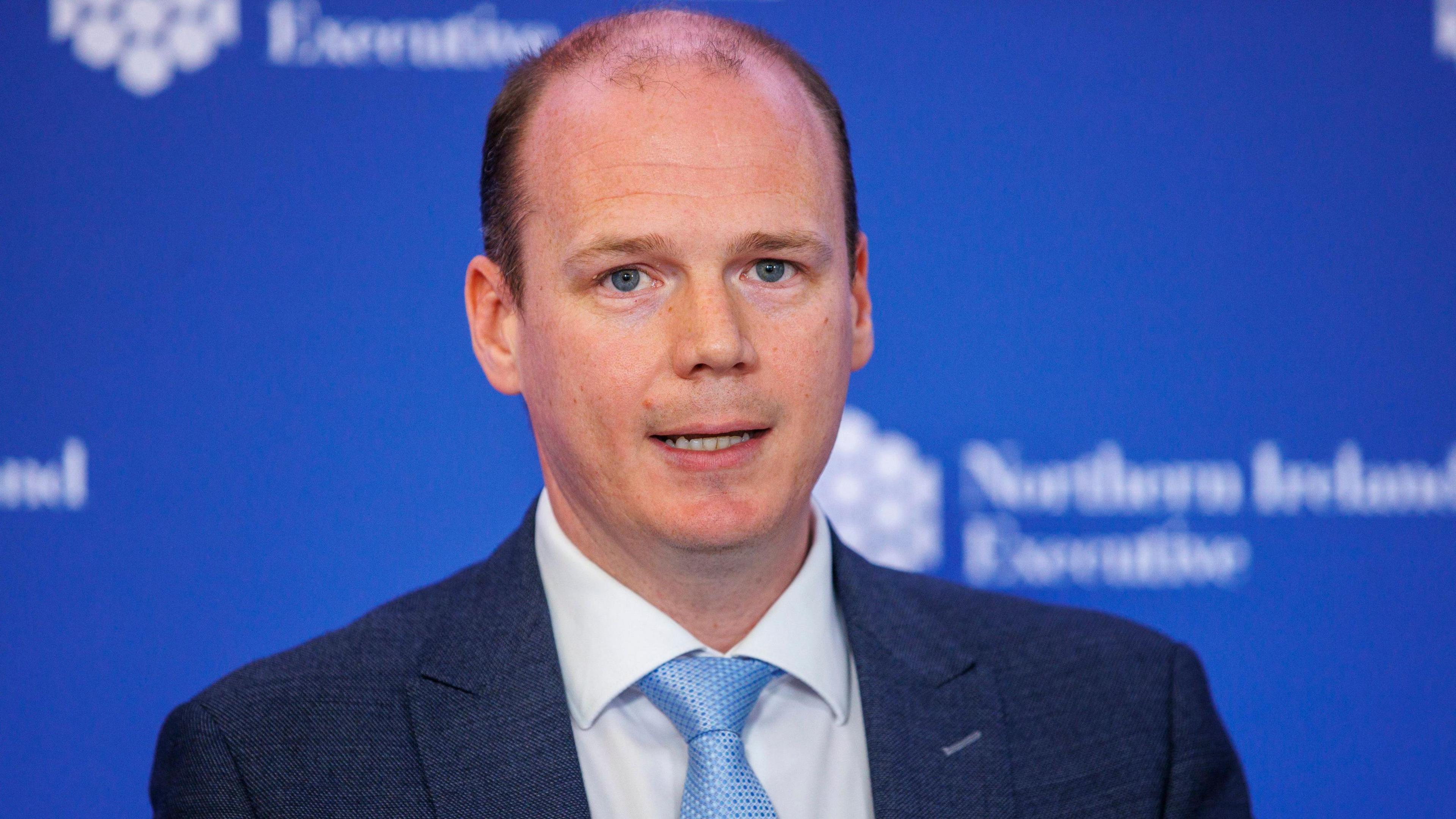 A man with short brown hair, wearing a navy suit jacket, white shirt and blue tie