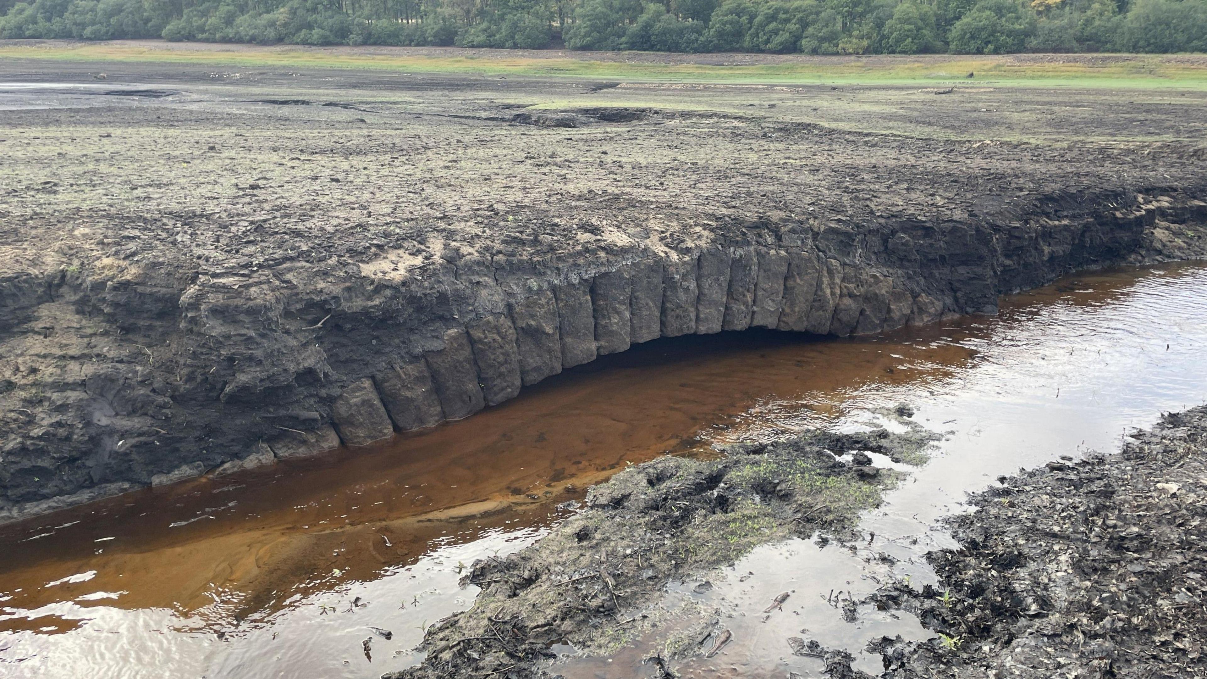 Some water flowing across what looks like a muddy field but is actually a nearly-empty reservoir. The water forms a stream which only looks a few inches deep. A stone arch is peeking out of the ground and shallow water by about a foot.