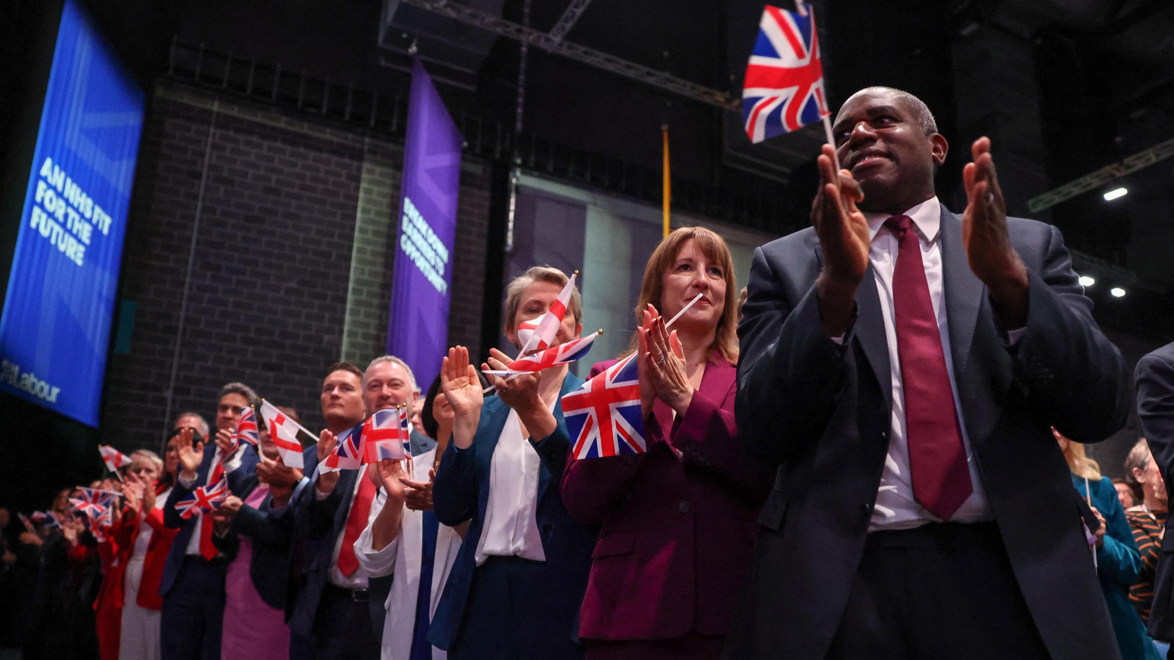Senior Labour ministers lined up clapping at the Labour Party conference, holding mini union jack flags.