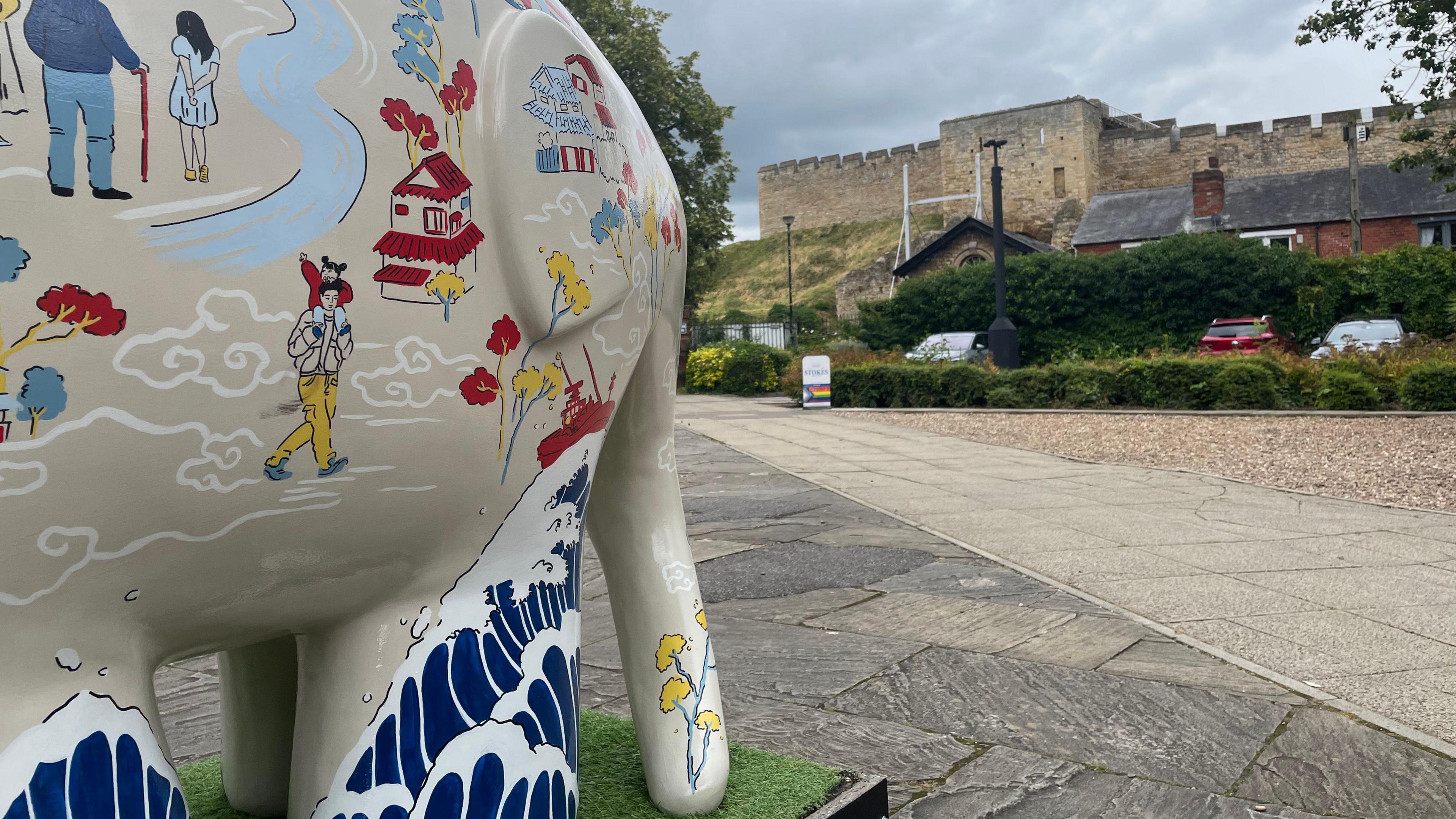 A cream-coloured Elmer the Elephant statue standing on a plinth looking towards the back of Lincoln Castle and has drawings of adults and children, with one child on the shoulders of a man.