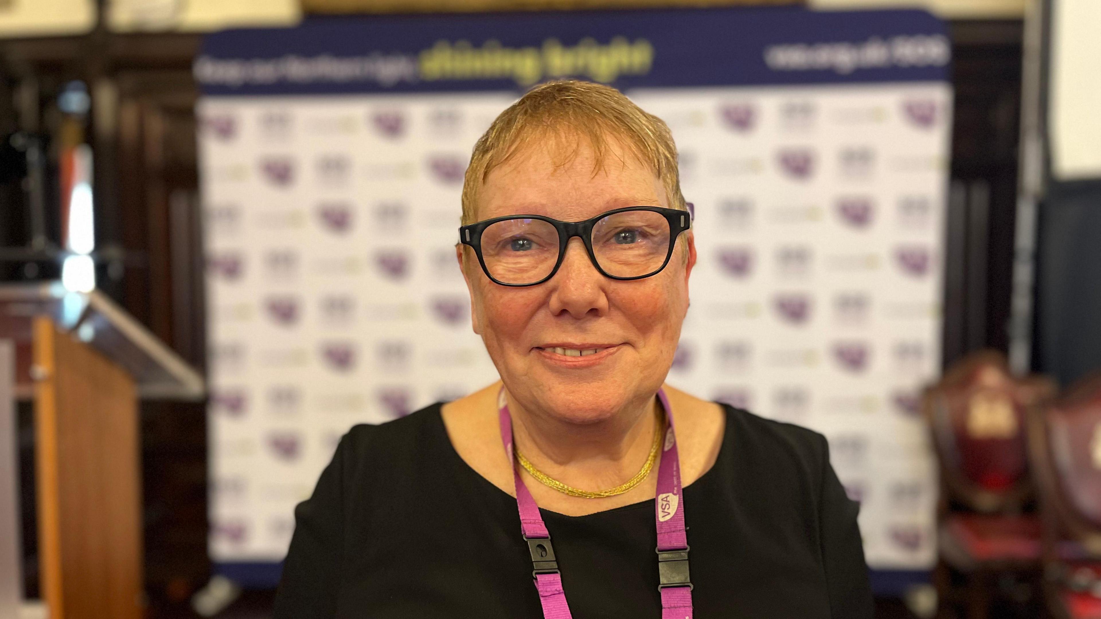 Sue Freeth. She has short hair and vision glasses with black frames is standing in the middle of the shot and looking into the camera. She is wearing black and has a lanyard on her neck with charity's logo. The promotional banner with charity's logo is in the background, that is blurred.