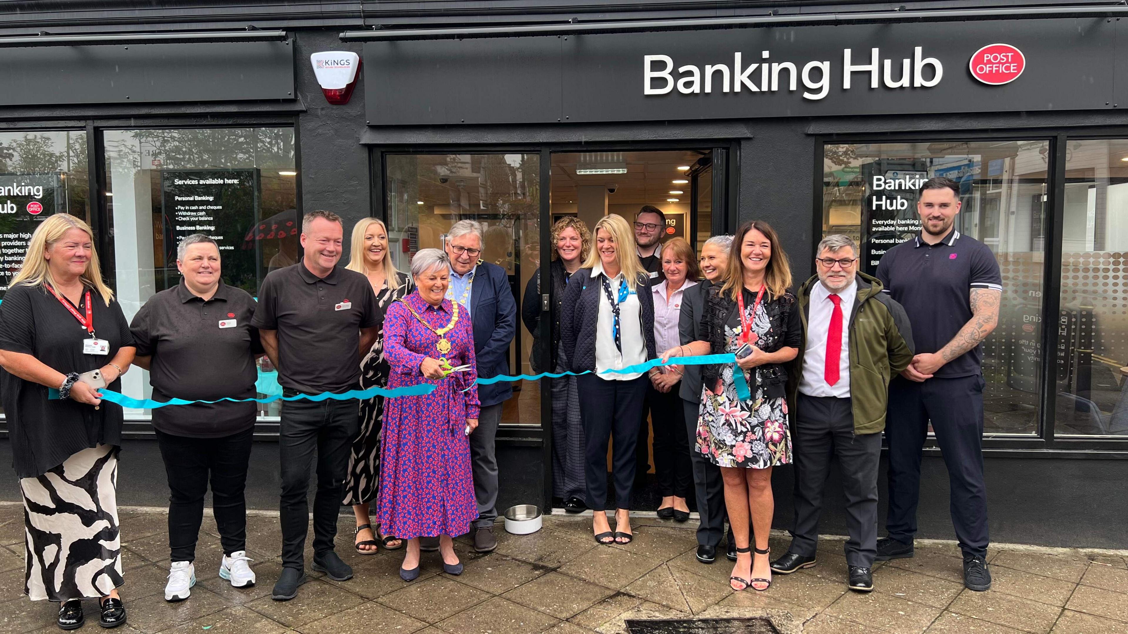 Mayor Julia Peggs stands in the middle of a line of 13 people outside the Banking Hub cutting a blue ribbon which has been cut but is yet to fall to the floor