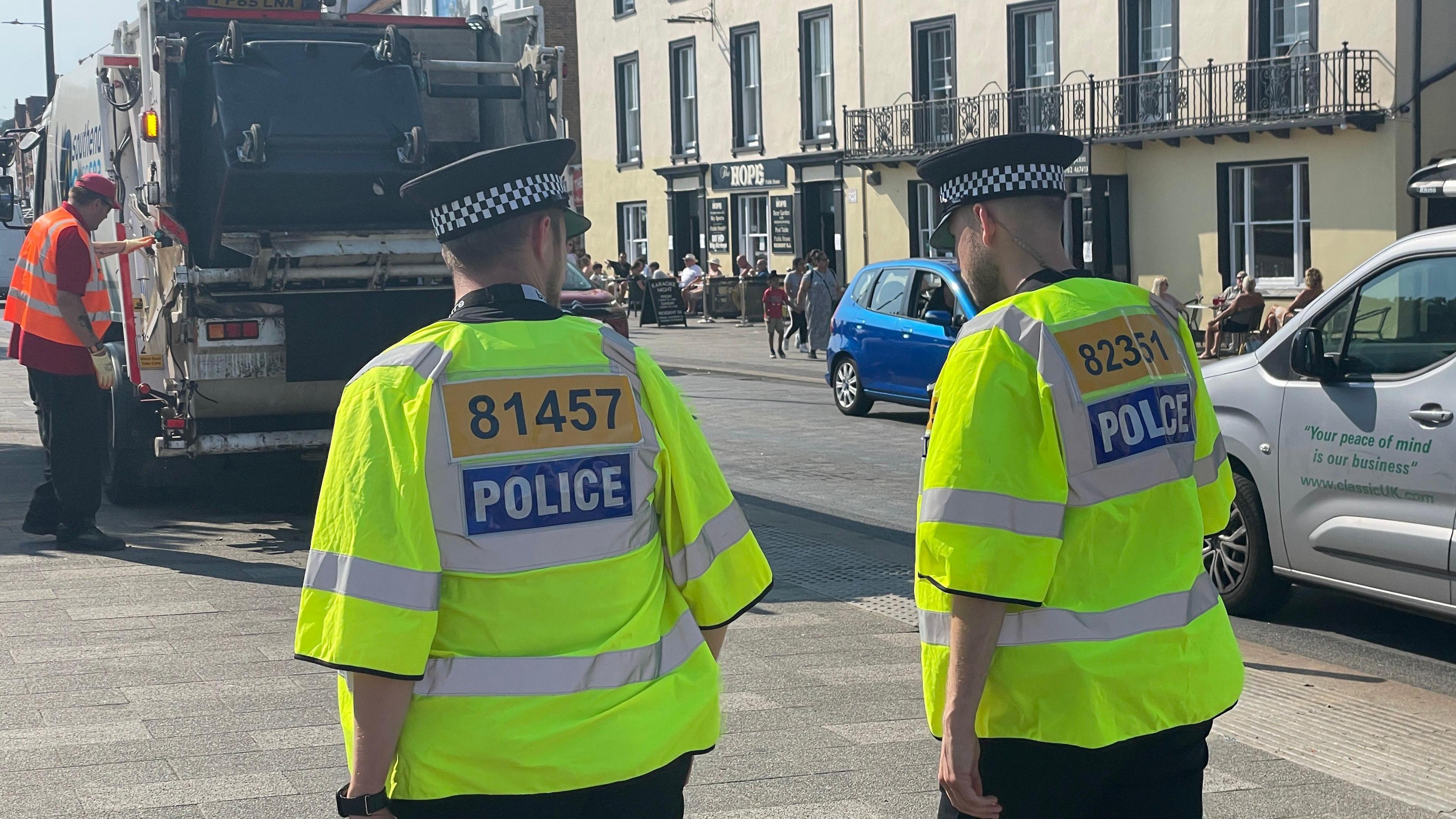 Two police officers wearing high vis jackets walking along  a street in Southend. Ahead of them is a bin lorry and a refuse worker, while cars and pedestrians can also be seen.