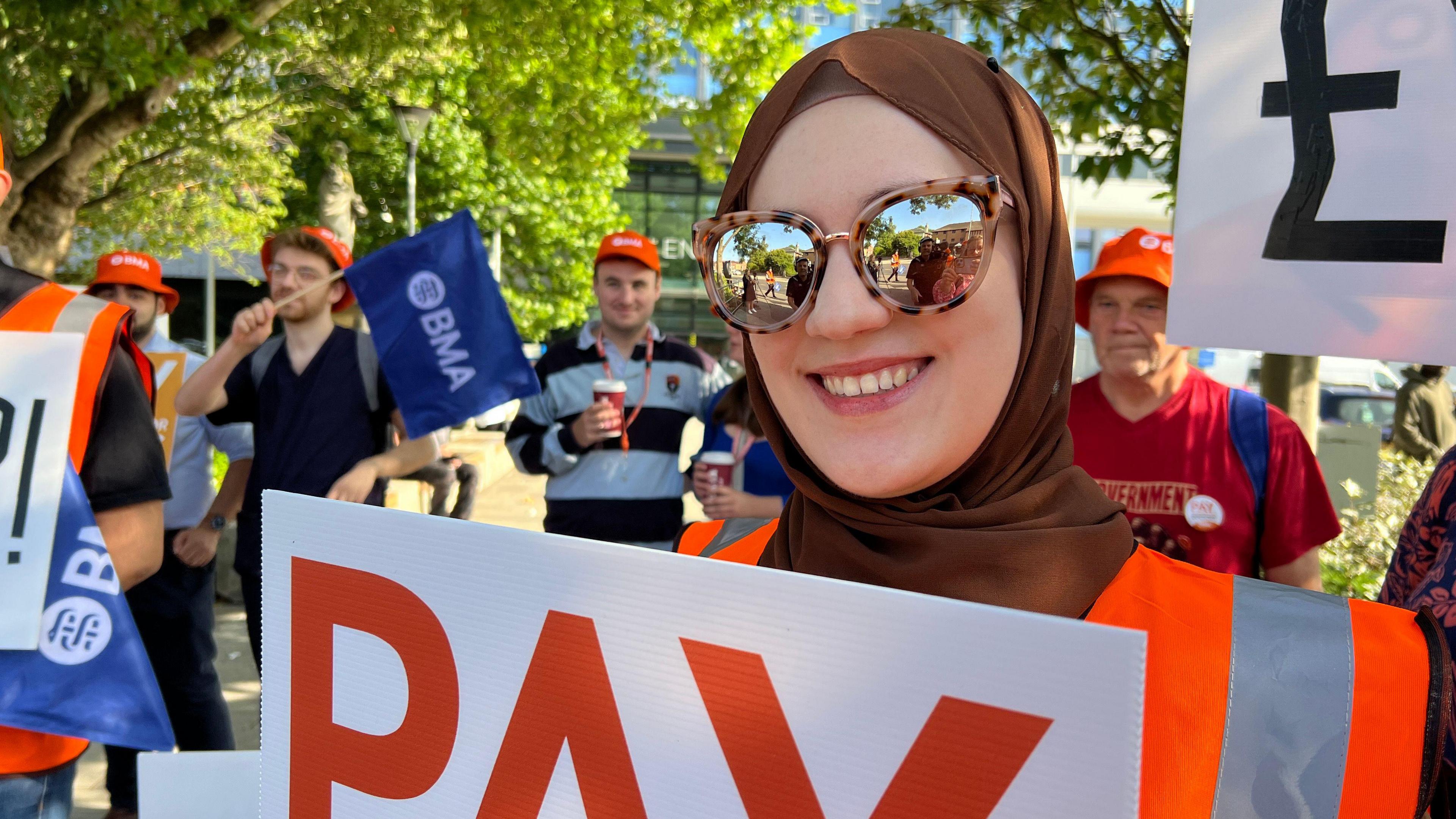 A woman wearing a brown headpiece with cheetah-print sunglasses, smiling and holding up a sign. She is wearing an orange hi-vis top. Behind her are several other people also holding up signs.
