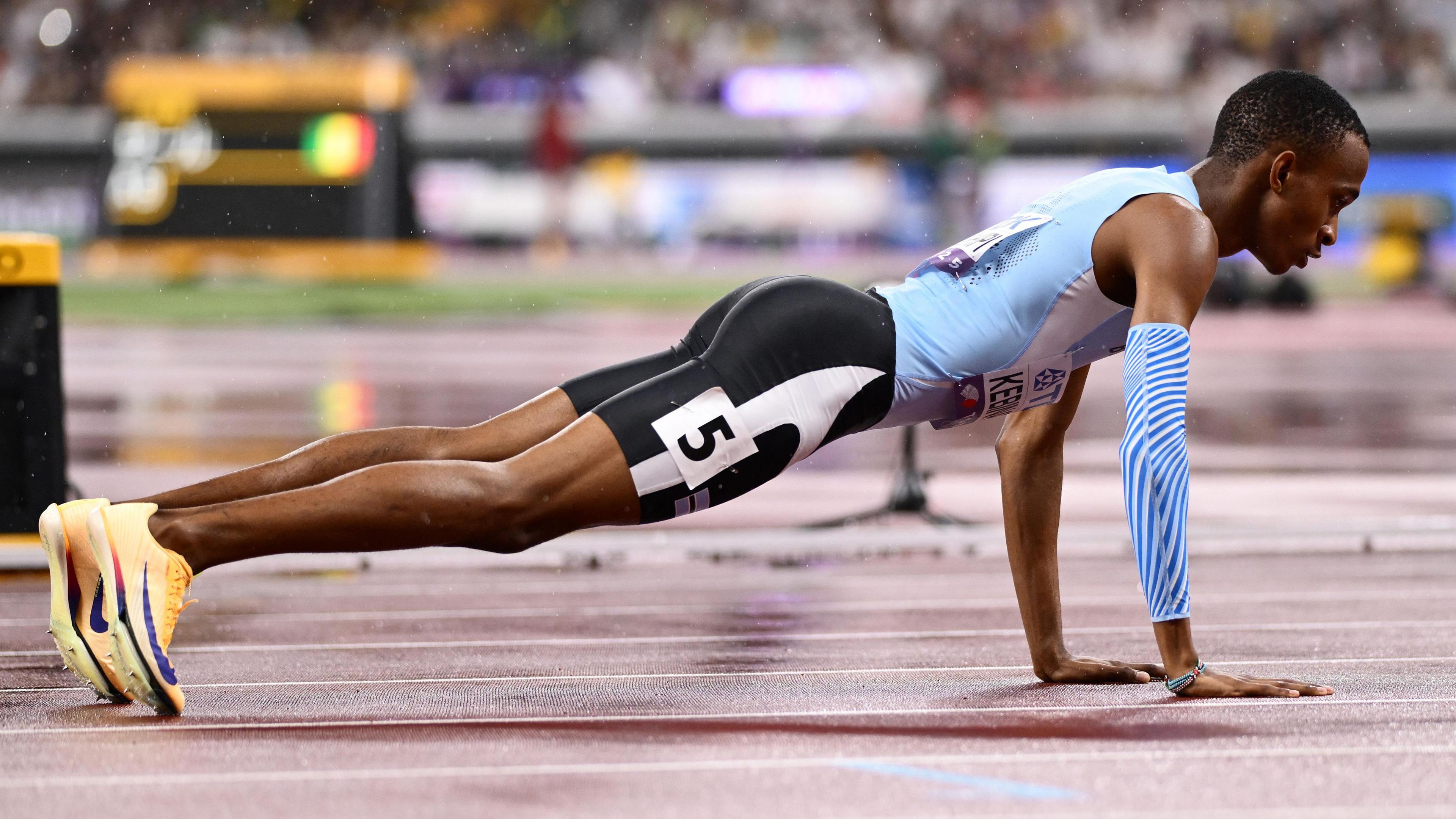 In a picture taken almost at track level, Collen Kebinatshipi, wearing a blue running vest, black shorts with the number five on the side, a blue running sleeve covering part of his right arm and orange shoes, is seen holding the top of a push-up pose on a damp running track. Blurred in the background are athletics scoreboards, advertising hoardings and the lower tiers of a crowd seen across the field of competition at the 2025 World Championships in Tokyo