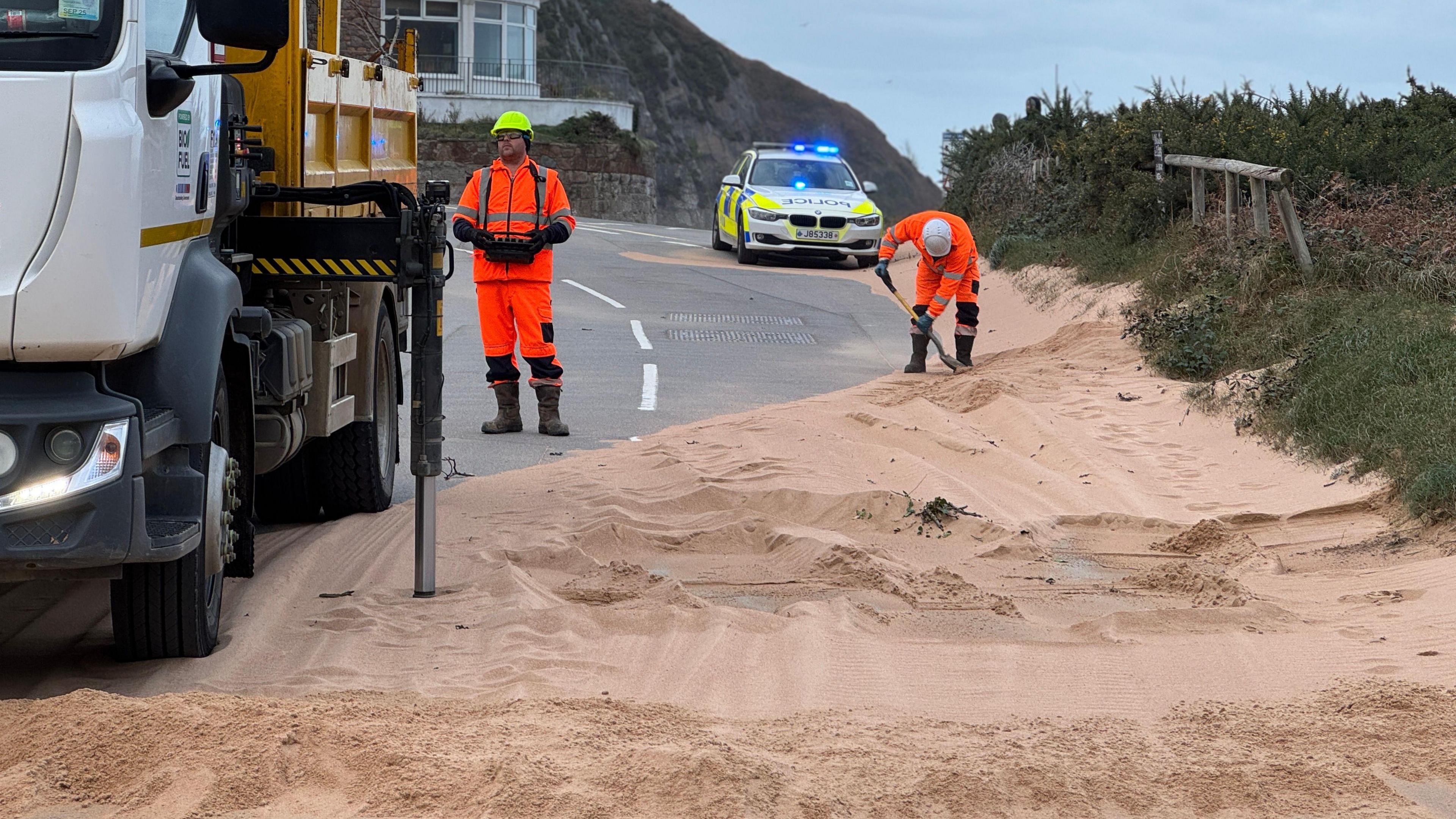 One man wearing an orange hi-viz uniform is shovelling sand which is piled up across the road and another is standing by a lorry. Behind them is a parked police car with its blue lights flashing. 