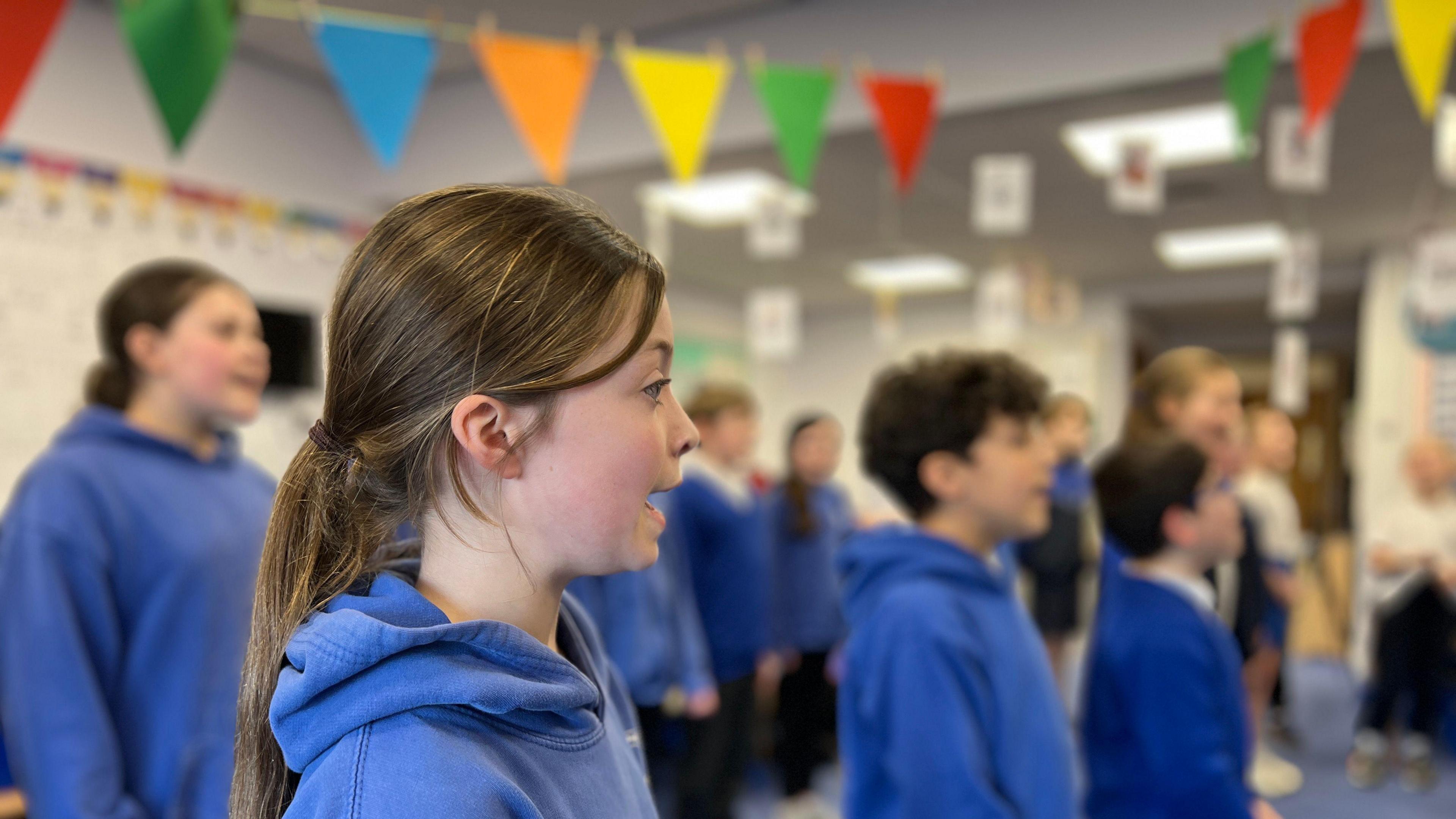 A group of children signing. A girl in the foreground with long light brown hair tied into a ponytail is in focus and the others are blurred behind her. The pupils are all wearing blue hoodies. 