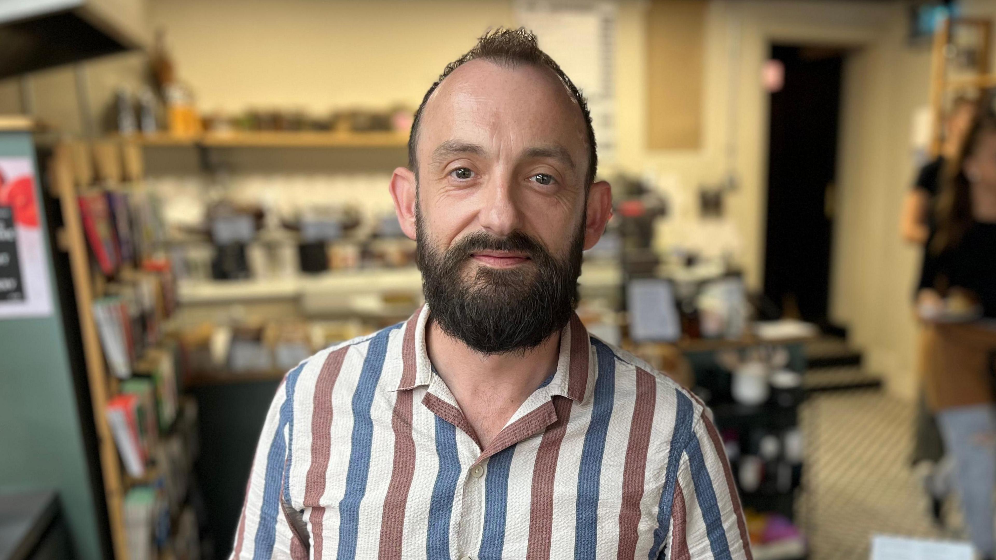 Alex Middleton from the shoulders up. He has brown hair and a brown beard. Behind him is the cafe out of focus.