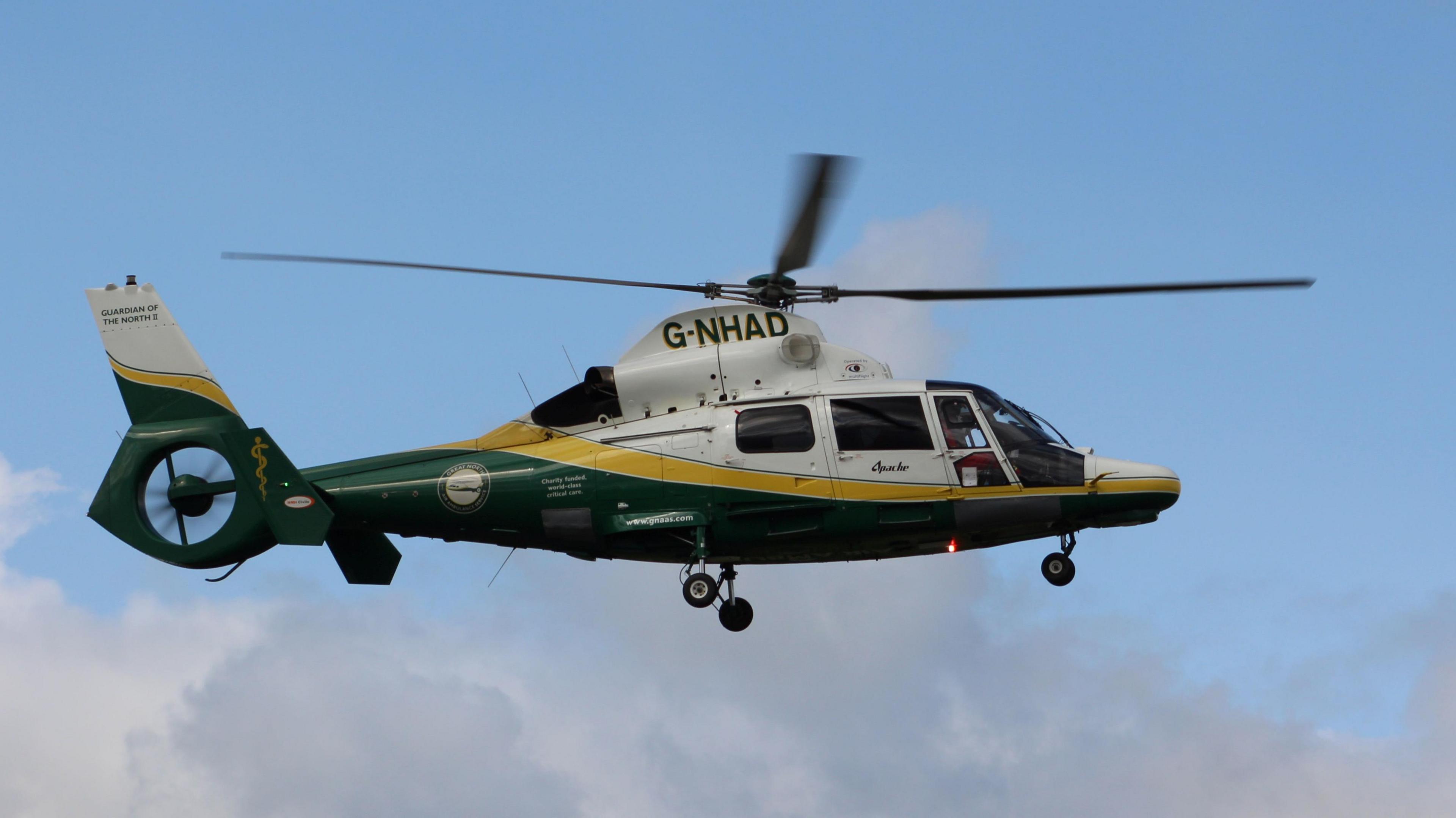 A GNAAS helicopter flying against a blue sky with a few fluffy clouds. The helicopter is white, yellow and green.