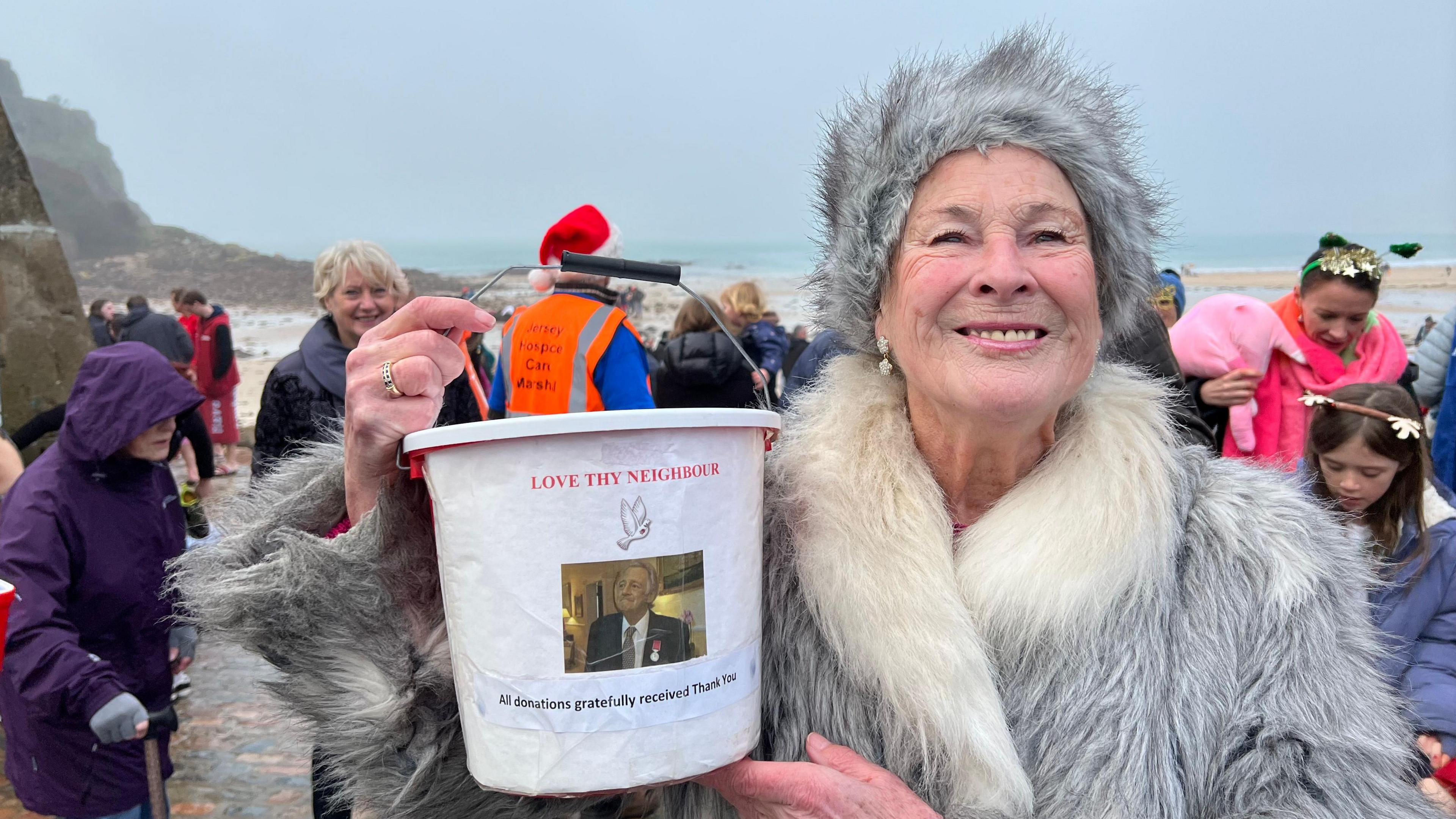 Ruth Curzon is an older woman wearing a furry hat and coat, she is holding a Love Thy Neighbour collection bucket. She is standing on the beach with swimmers behind her.