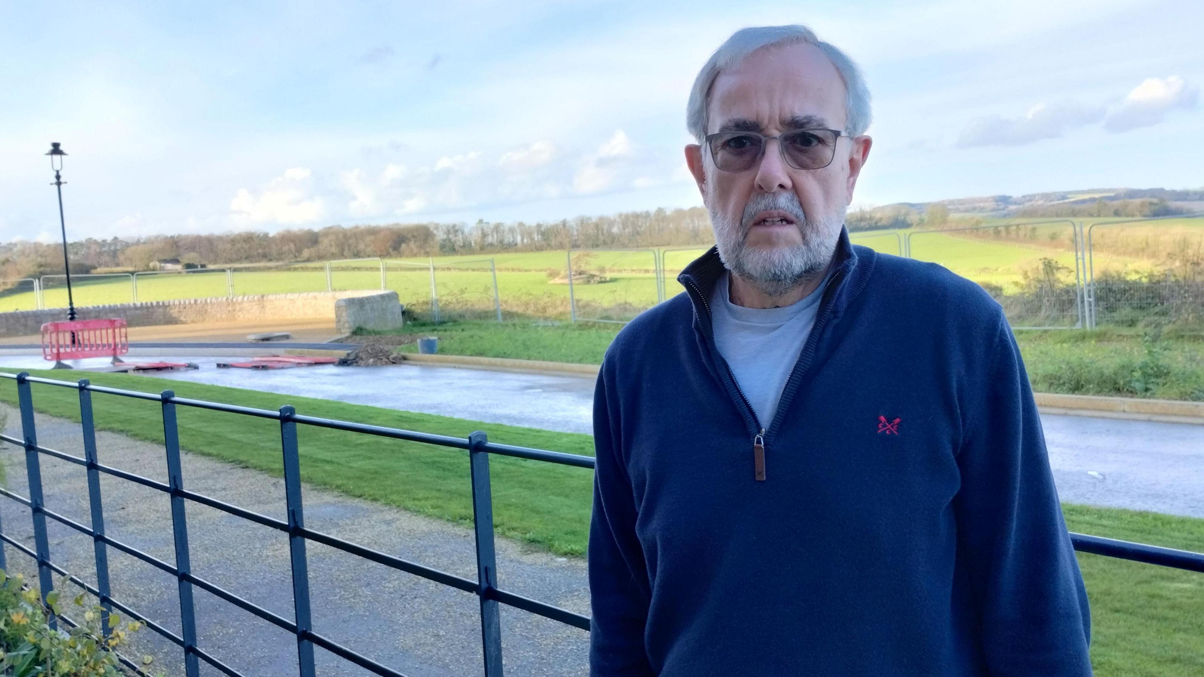 Andrew Cook stands in Peninsula Way. He has glasses, grey hair and a beard and wears a navy blue fleece. On the far side of the road is a linked wire fence bordering fields.