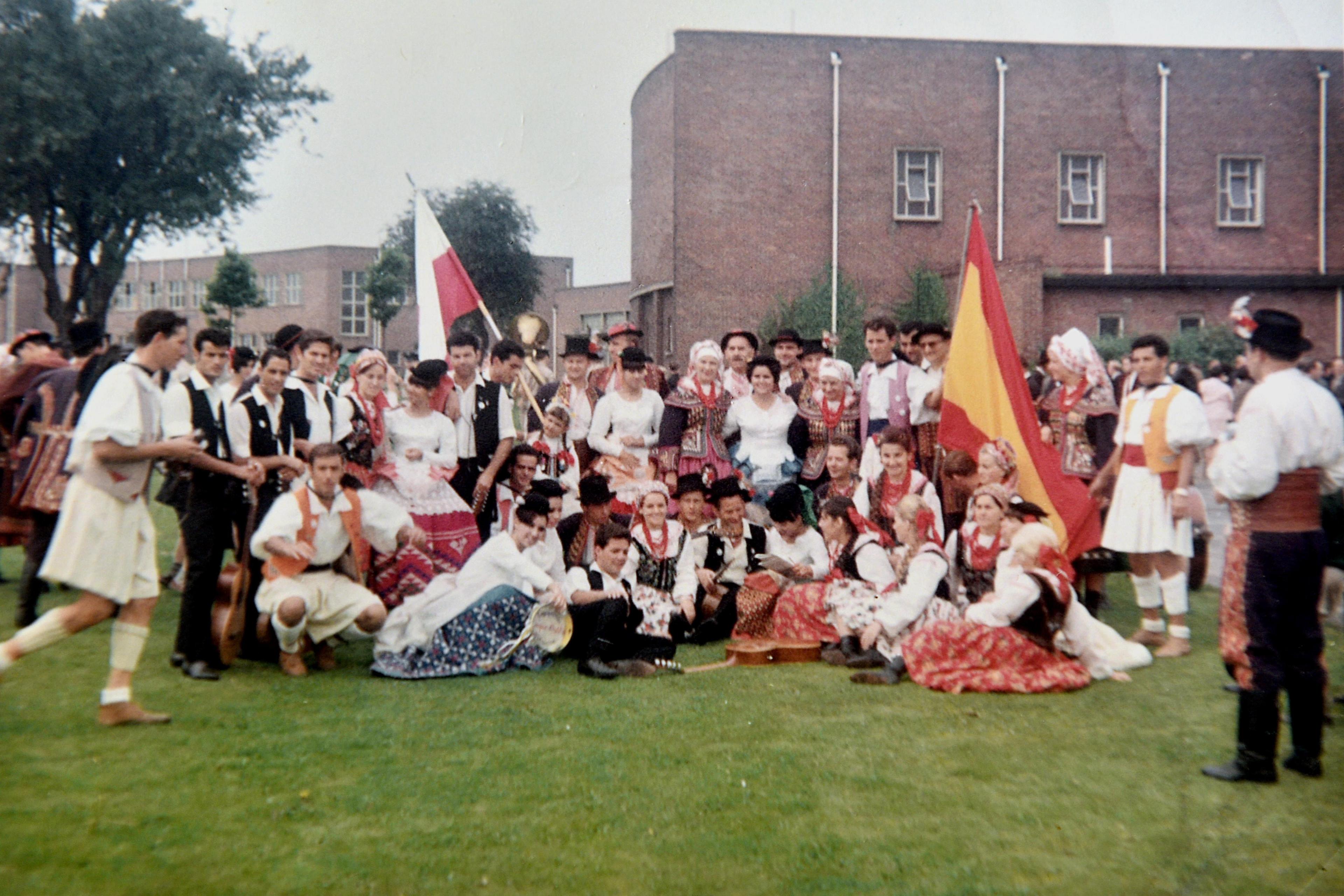 A big crowd of people sitting on grass. One person is holding a Spanish flag, the other is holding a Polish flag. Everyone is in traditional folk clothing.