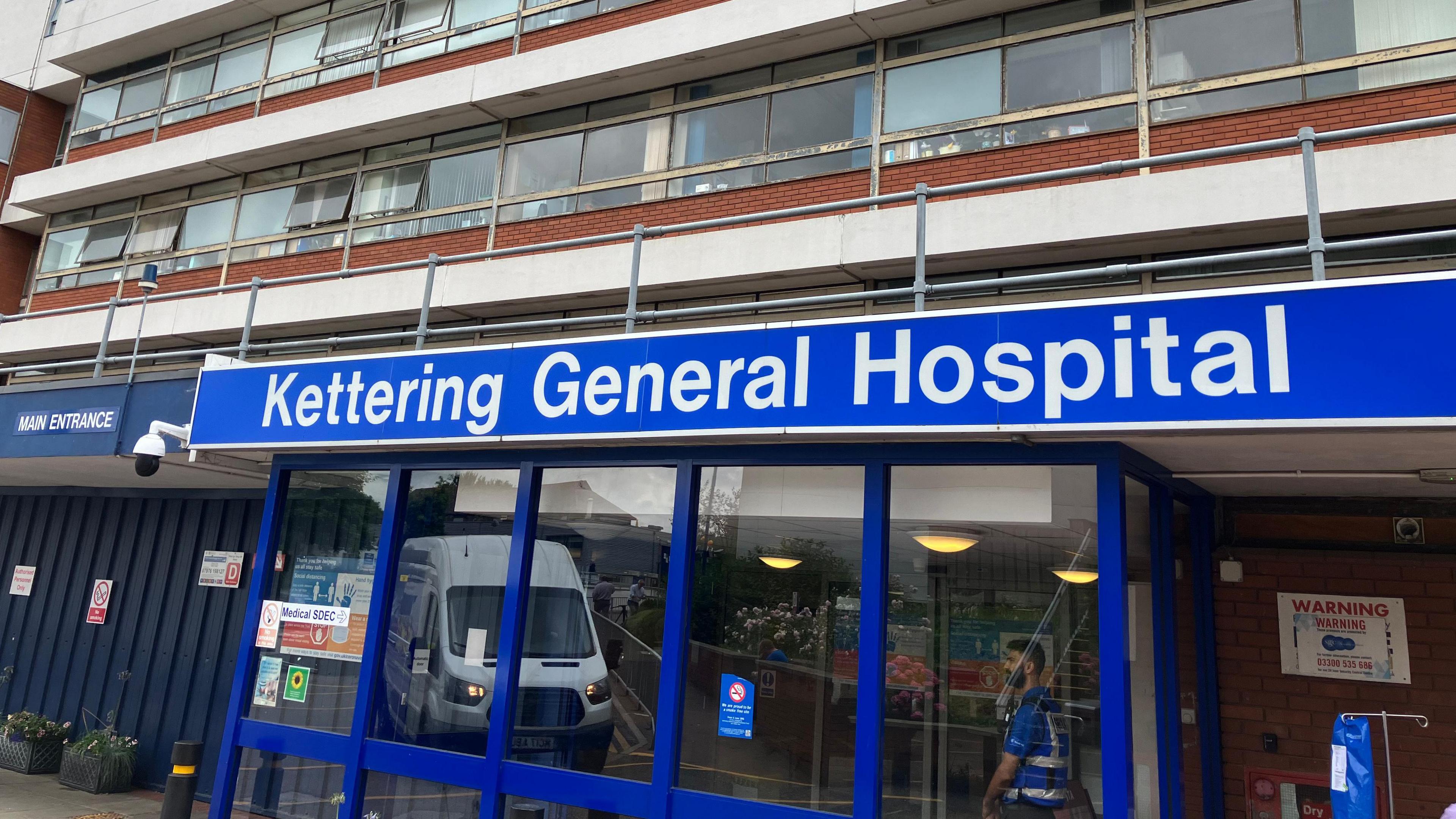 The outside of Kettering General Hospital, a blue glass-fronted entrance with "Kettering General Hospital" in large letters. 