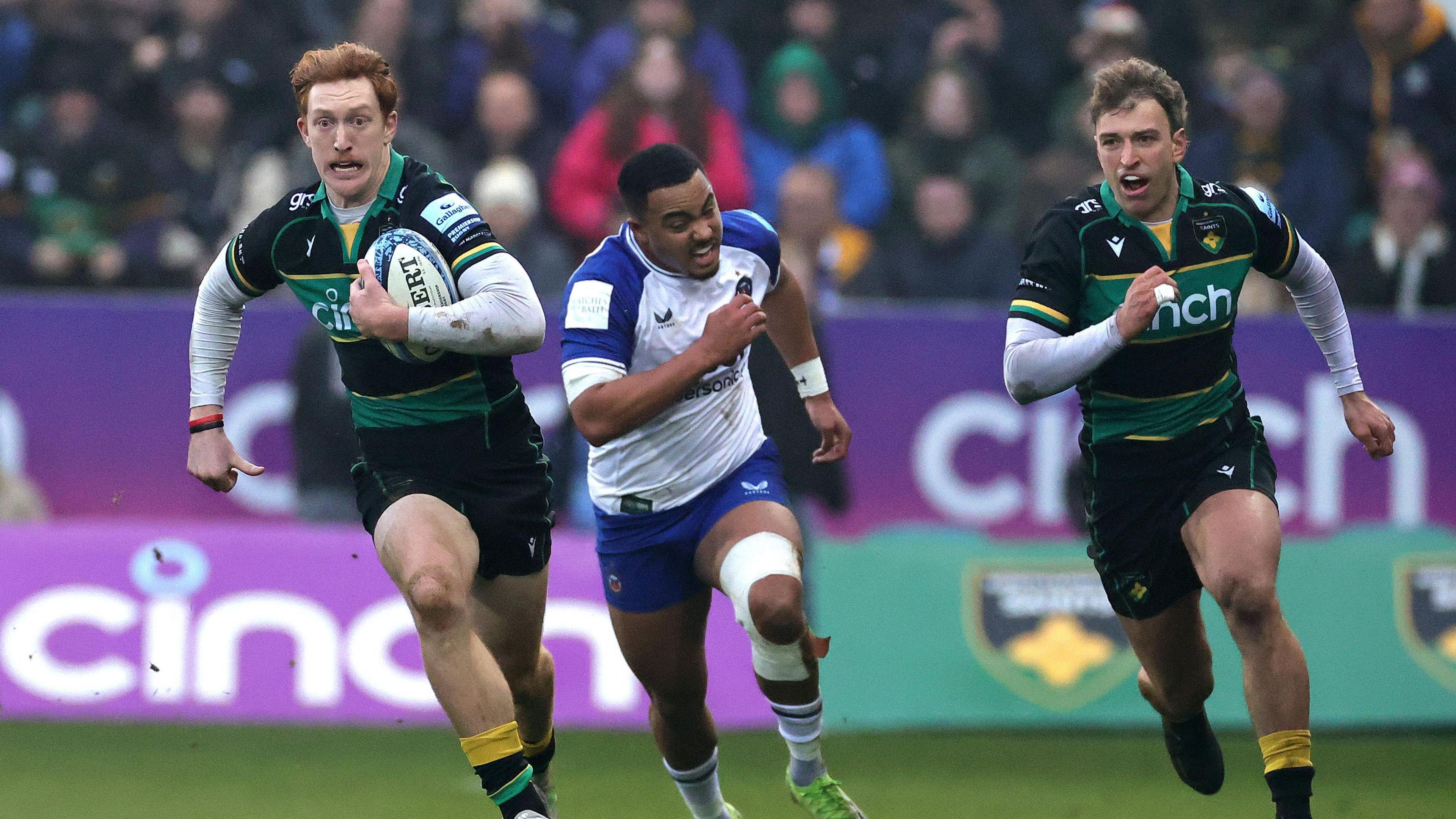 man running with rugby ball wearing green/yellow and black strip. two men in his wake, one shouting, wearing green/yellow and black strip and the other in white and blue - crowd out of focus in background