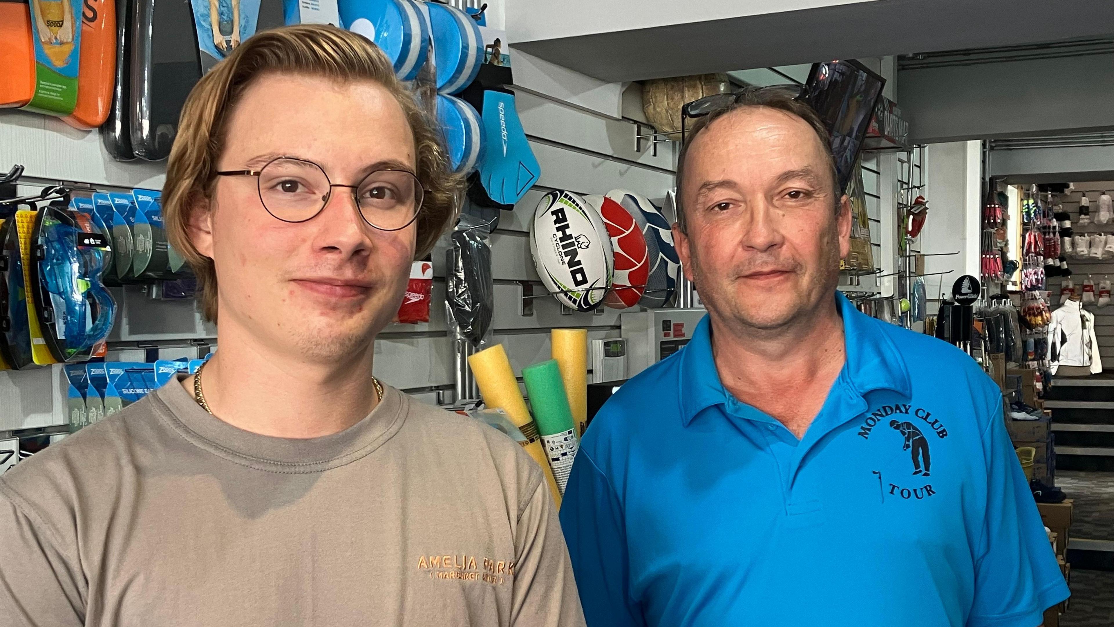 Two people stand next to each other, one wearing glasses and the other a blue t-shirt. They are surrounded by sports equipment and rugby balls. 