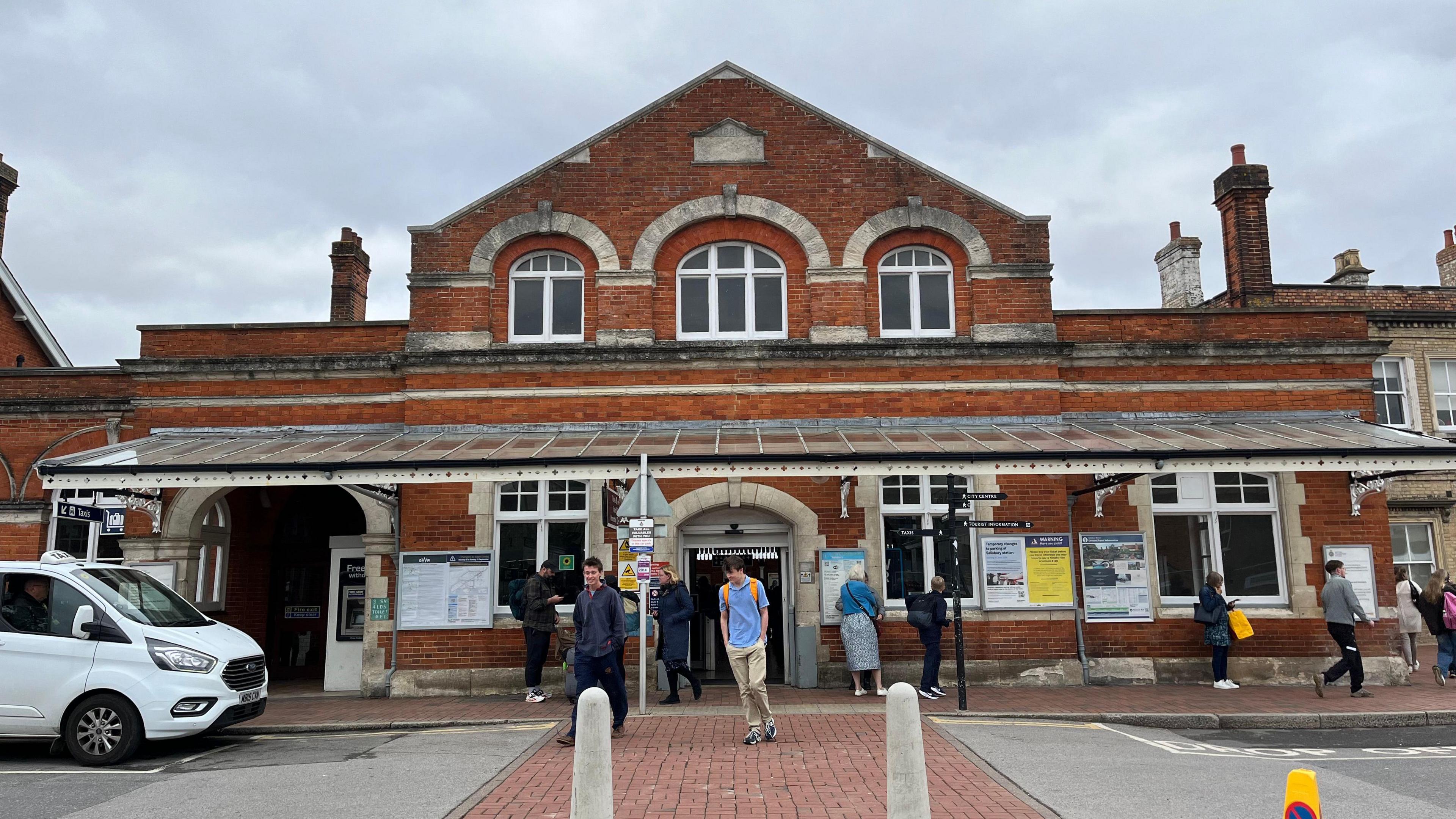 The front of Salisbury Railway Station with a Victorian-style red brick entrance and passengers and a white taxi visible