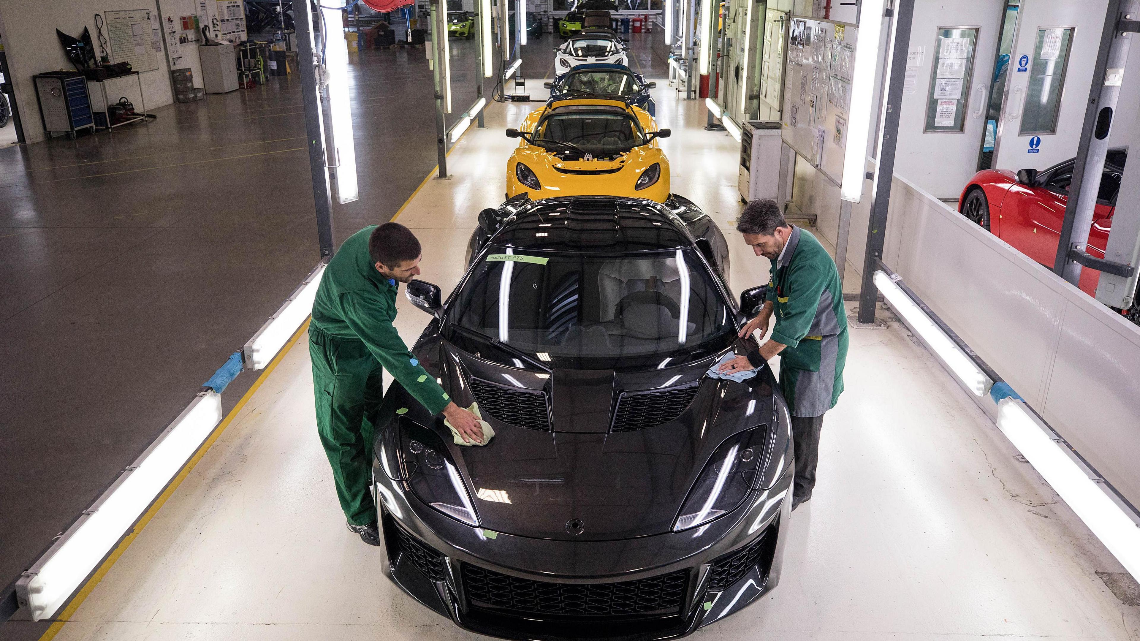 Two workers wearing green overalls polish a sports car at the front of a line of cars on a production line in a factory