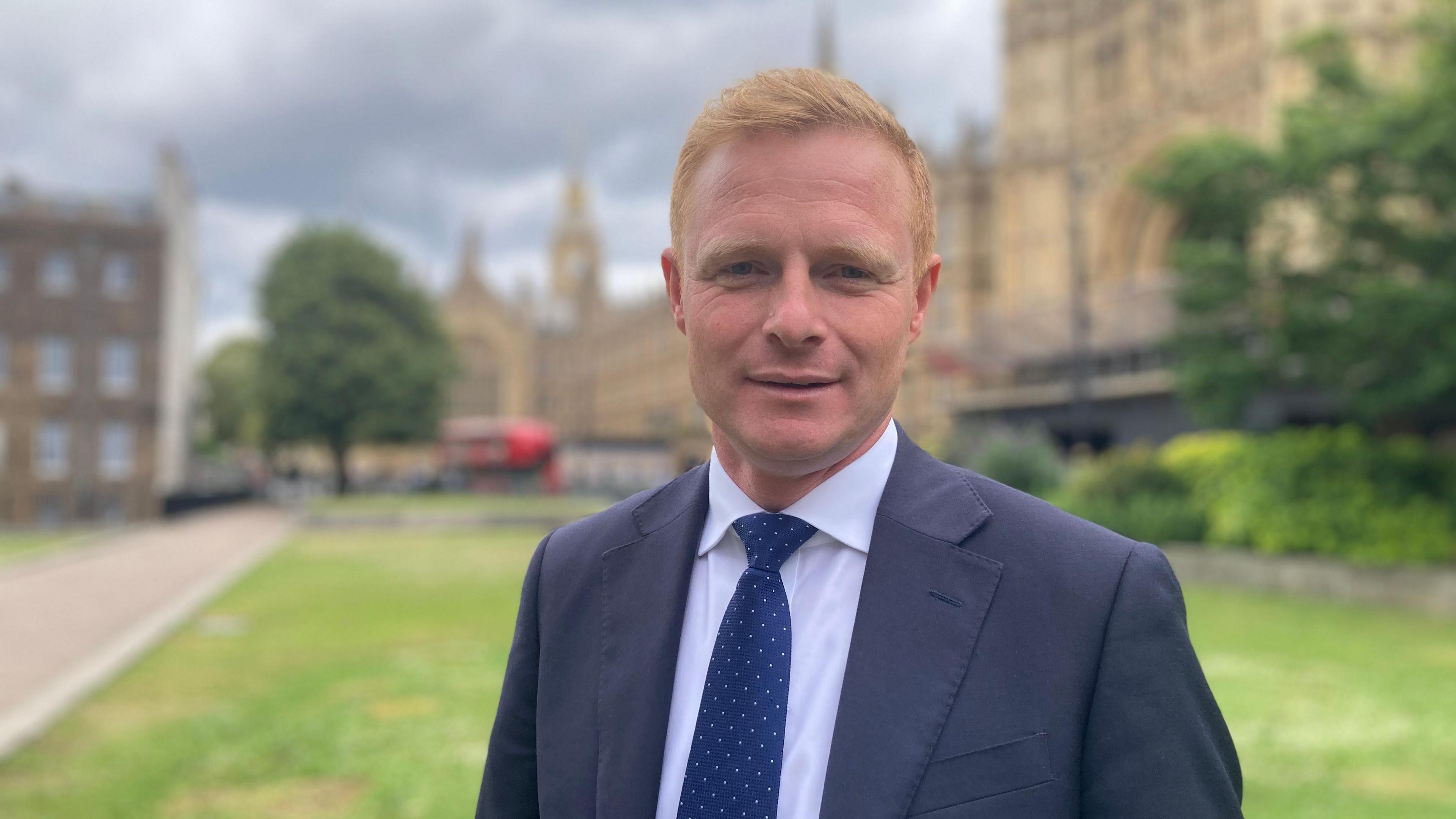 Robbie Moore, a man wearing a navy suit pictured on College Green near the House of Commons. He has red hair and is wearing a blue tie with white spots.